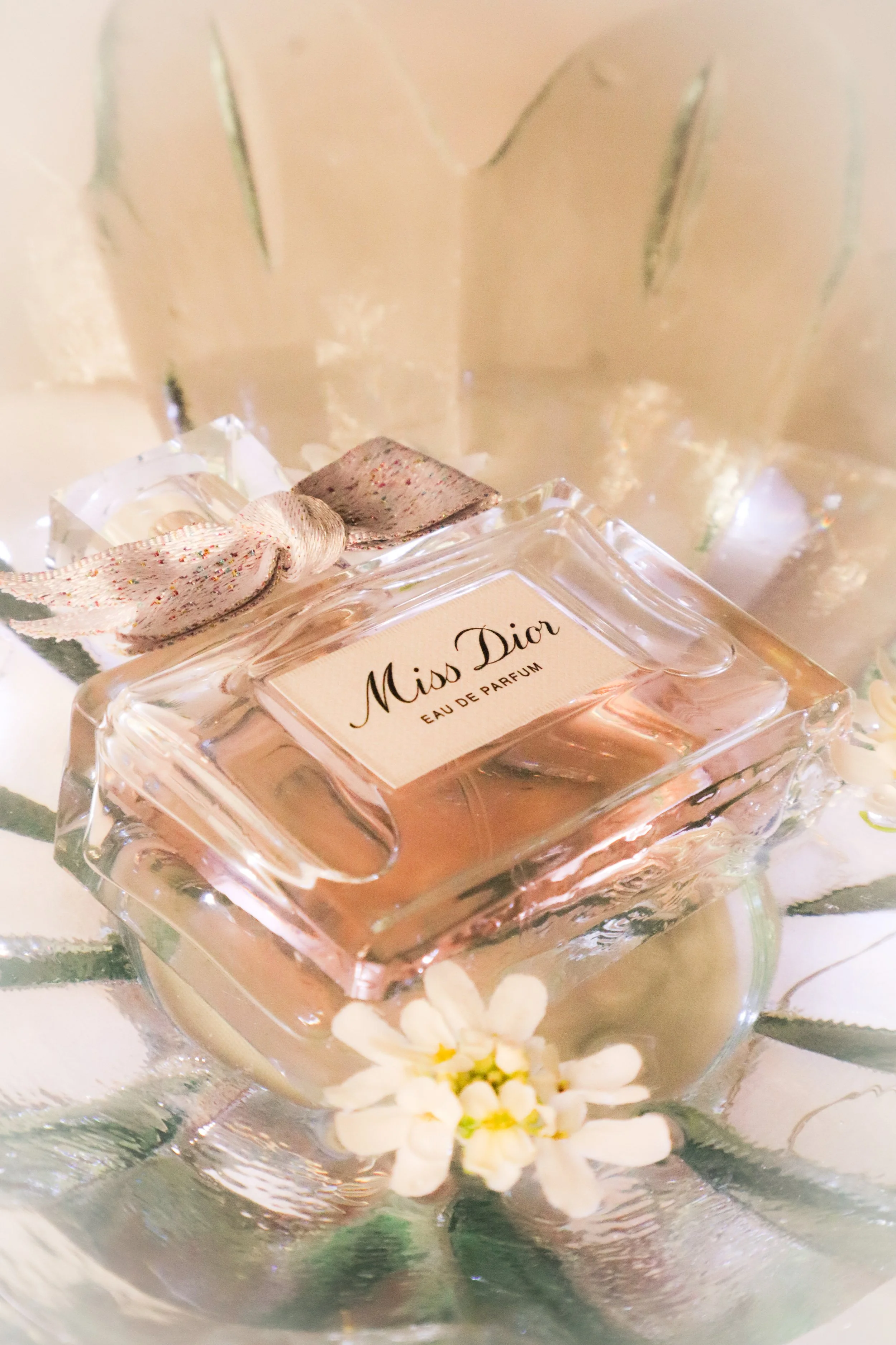 Un flacon de parfum Miss Dior Dans un récipient en verre avec une petite fleur blanche en premier plan.