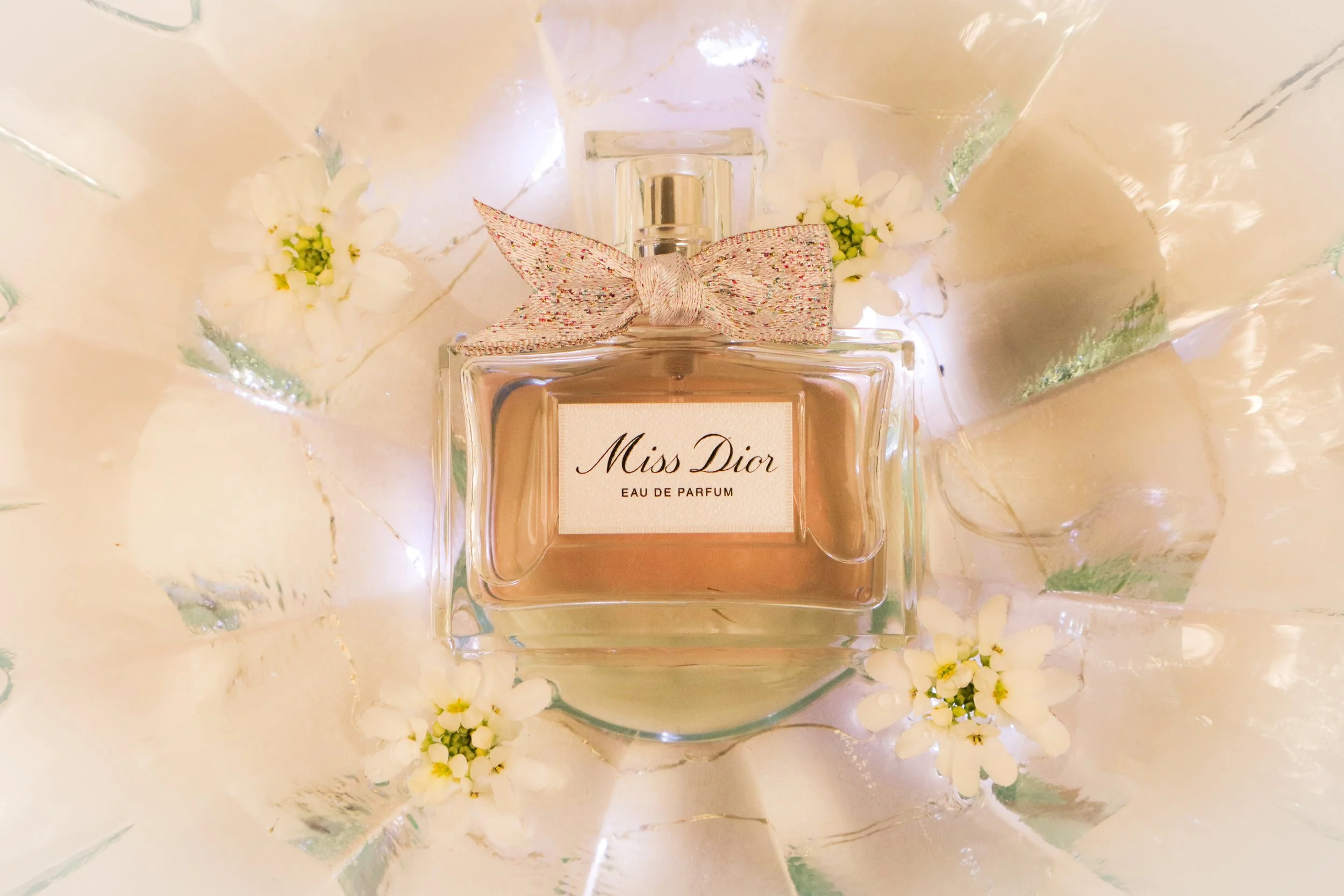 Flacon de parfum Miss Dior entouré de petites fleurs blanches sur un fond en miroir