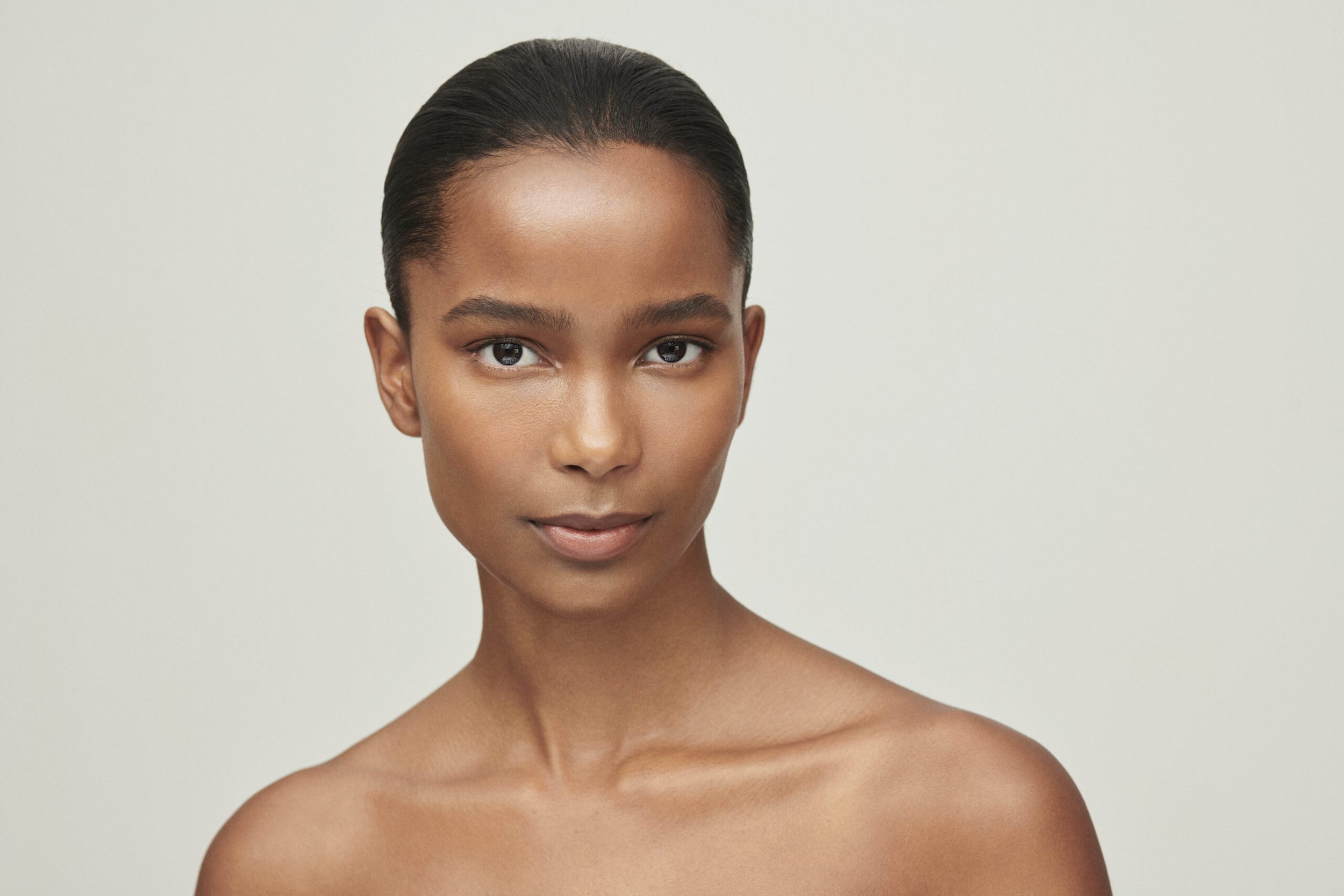 Close-up of a woman with dark skin and black hair styled back, looking directly at the camera with a neutral expression, against a plain light background.