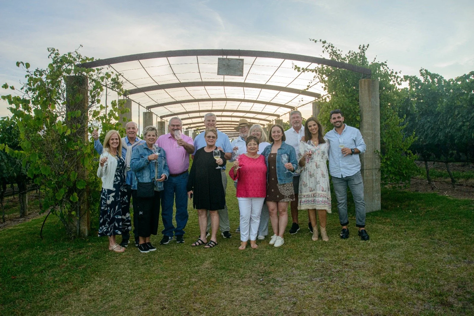 Group of people celebrating with drinks outdoors in a vineyard at sunset.