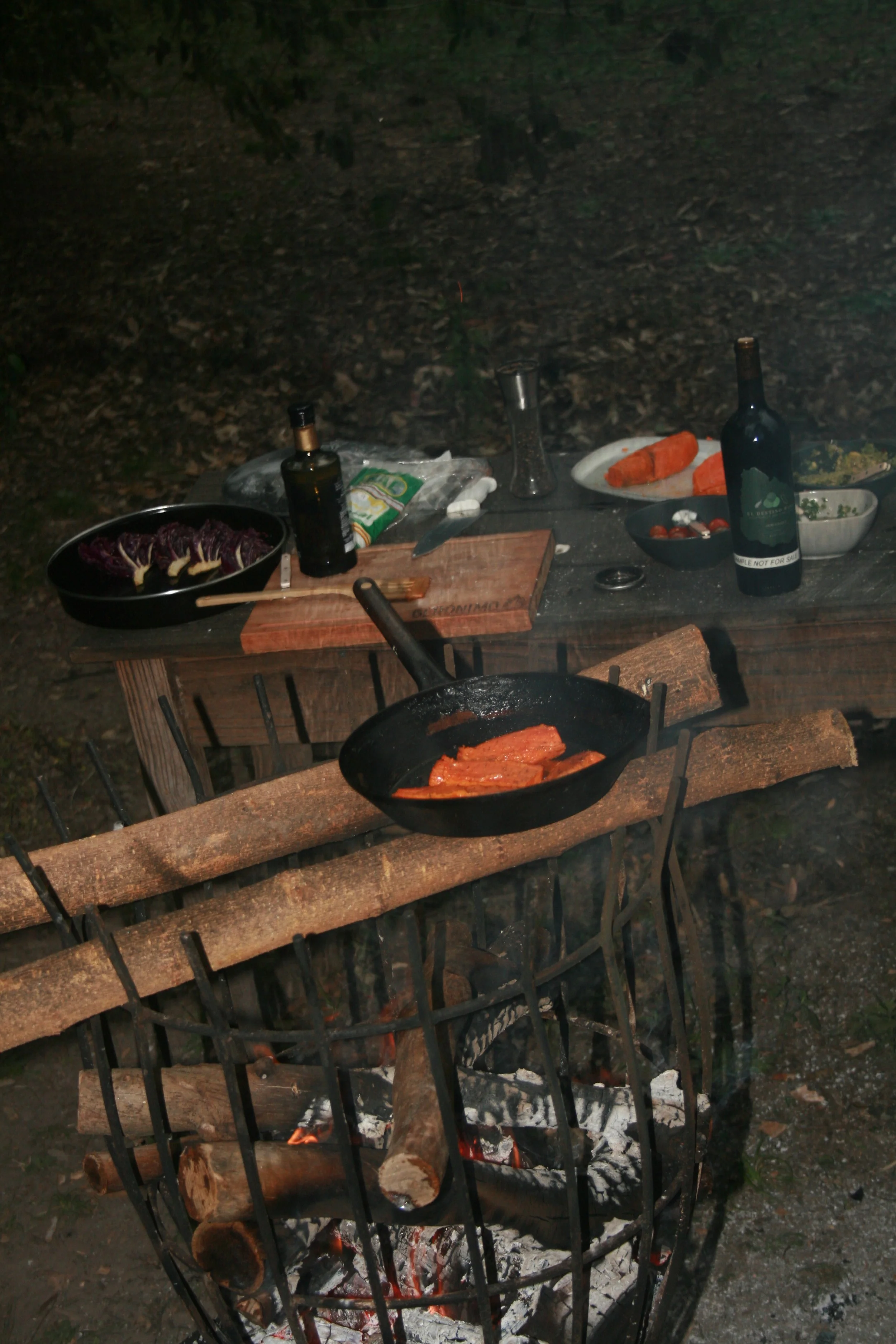 Outdoor campsite cooking scene with a grill over a campfire, featuring grilled salmon fillets, various vegetables, bottles of drinks, and plates of food on a wooden table in a forest setting at night.