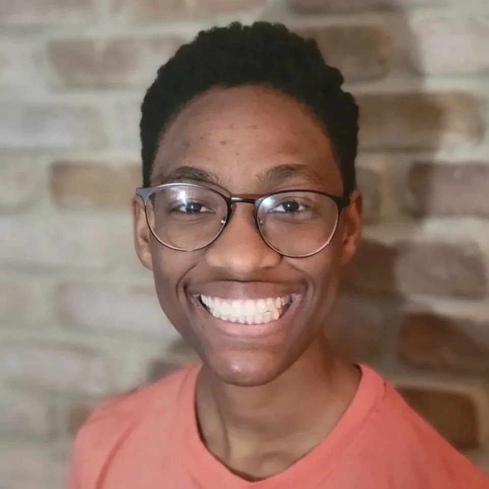 Young man smiling with glasses, wearing an orange shirt, against a brick wall background.