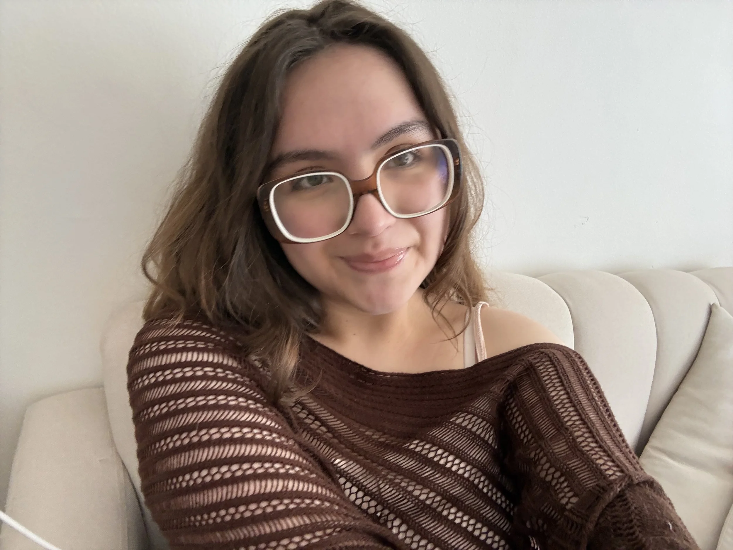 A woman with long brown hair and glasses smiling while sitting on a white couch, wearing a brown patterned long-sleeve top, against a plain white wall background.