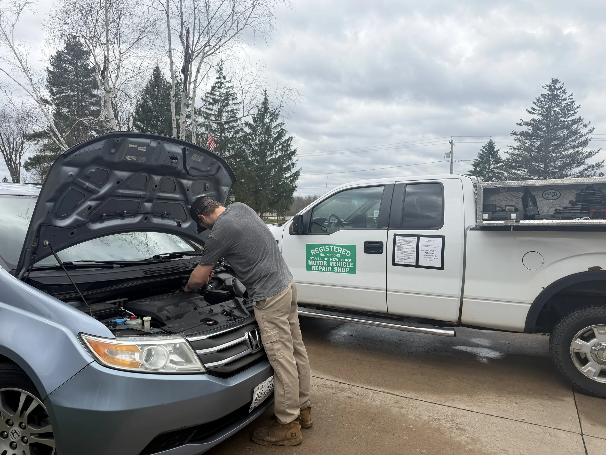 A man inspecting the engine under the open hood of a silver Honda sedan parked outdoors. Beside him is a white pickup truck with a green sign indicating it's a registered vehicle repair shop.
