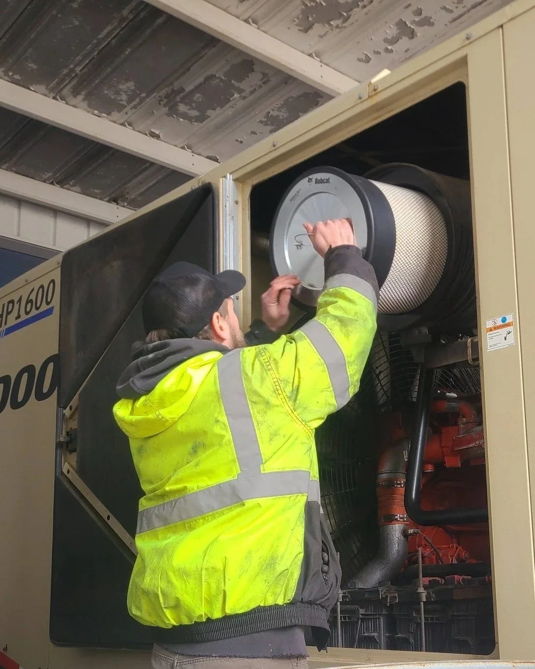 A worker in a high-visibility yellow jacket and black cap is inspecting or repairing a large industrial machine, possibly a generator, inside a metal structure.