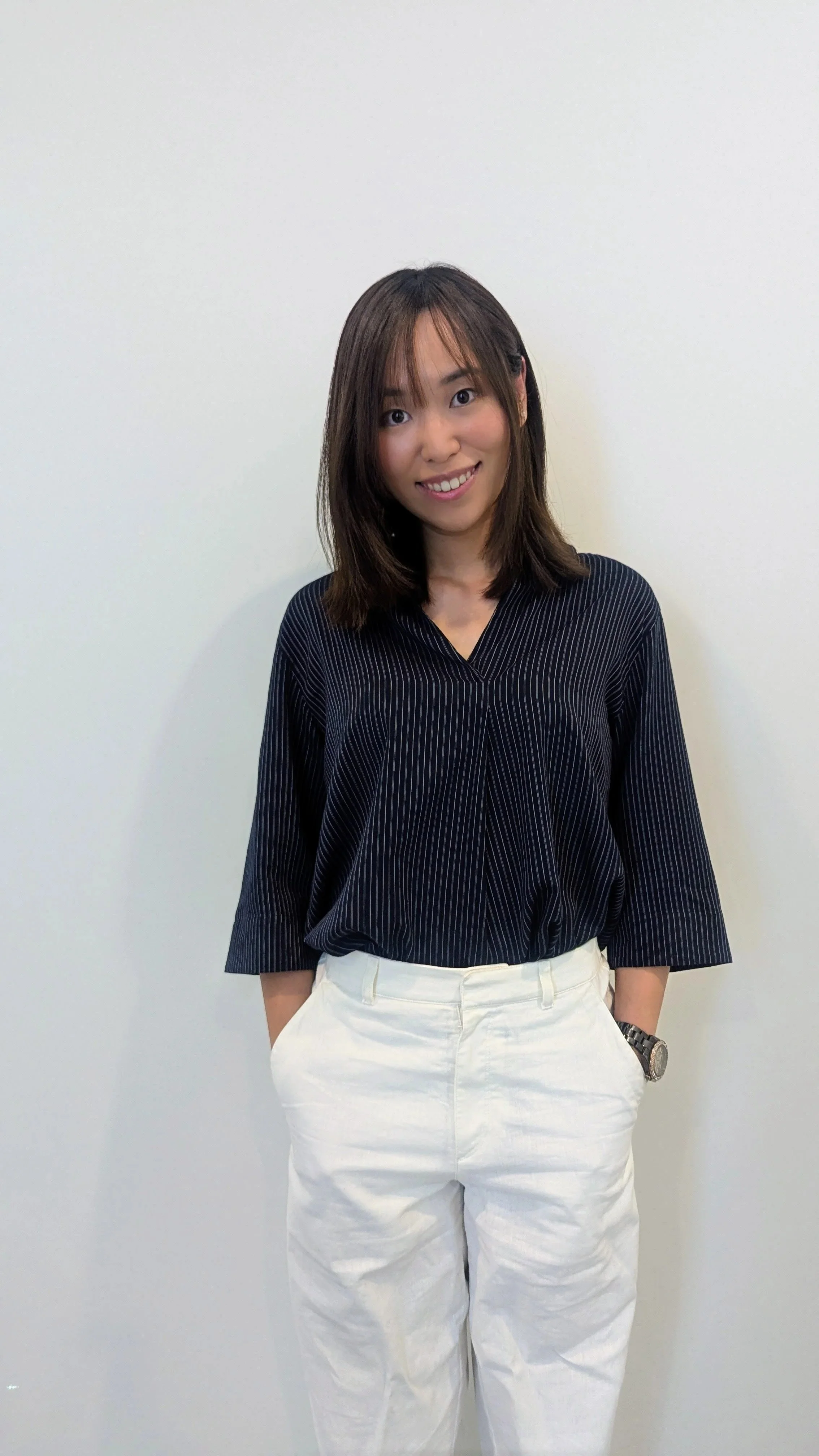 A woman with shoulder-length brown hair wearing a black and white pinstripe top and white pants standing against a plain white wall.