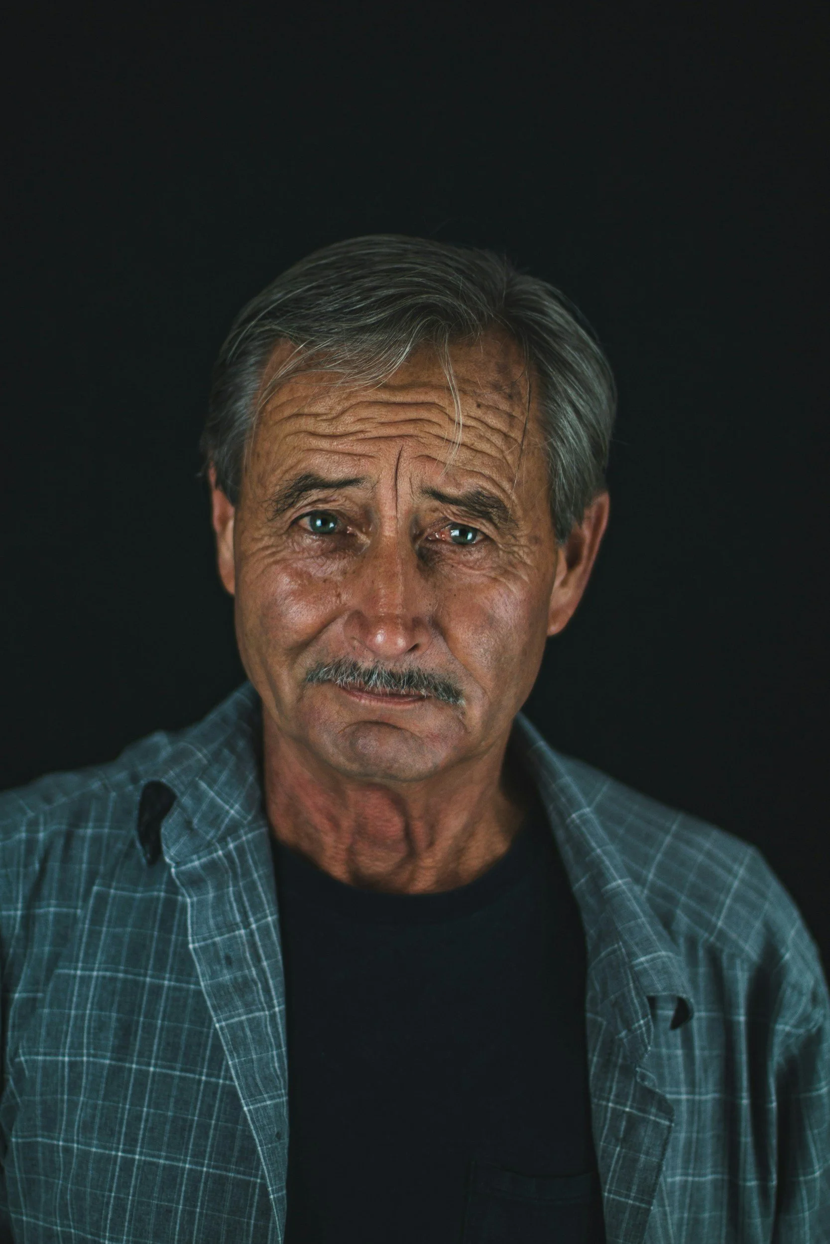 Close-up of an elderly man with gray hair, wearing a black T-shirt and a blue plaid shirt, against a black background.