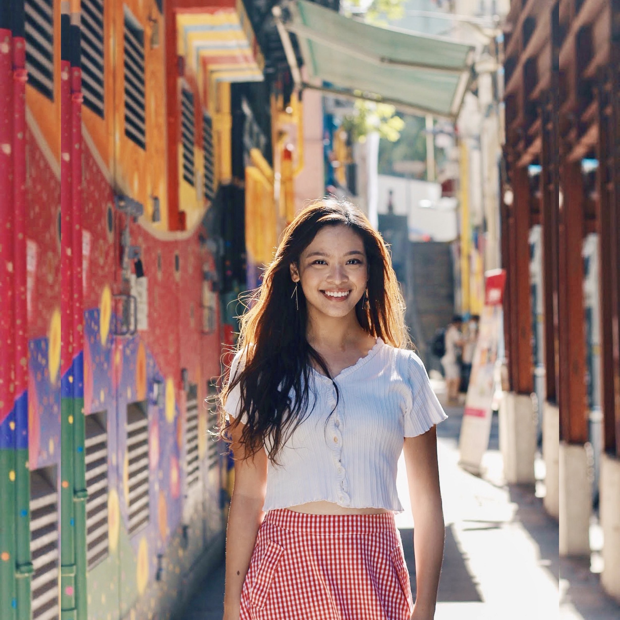 A young woman smiling outdoors standing in front of colorful fenced railing, wearing a white cropped top with buttons and a red checkered skirt, with sunny weather and blurred background.