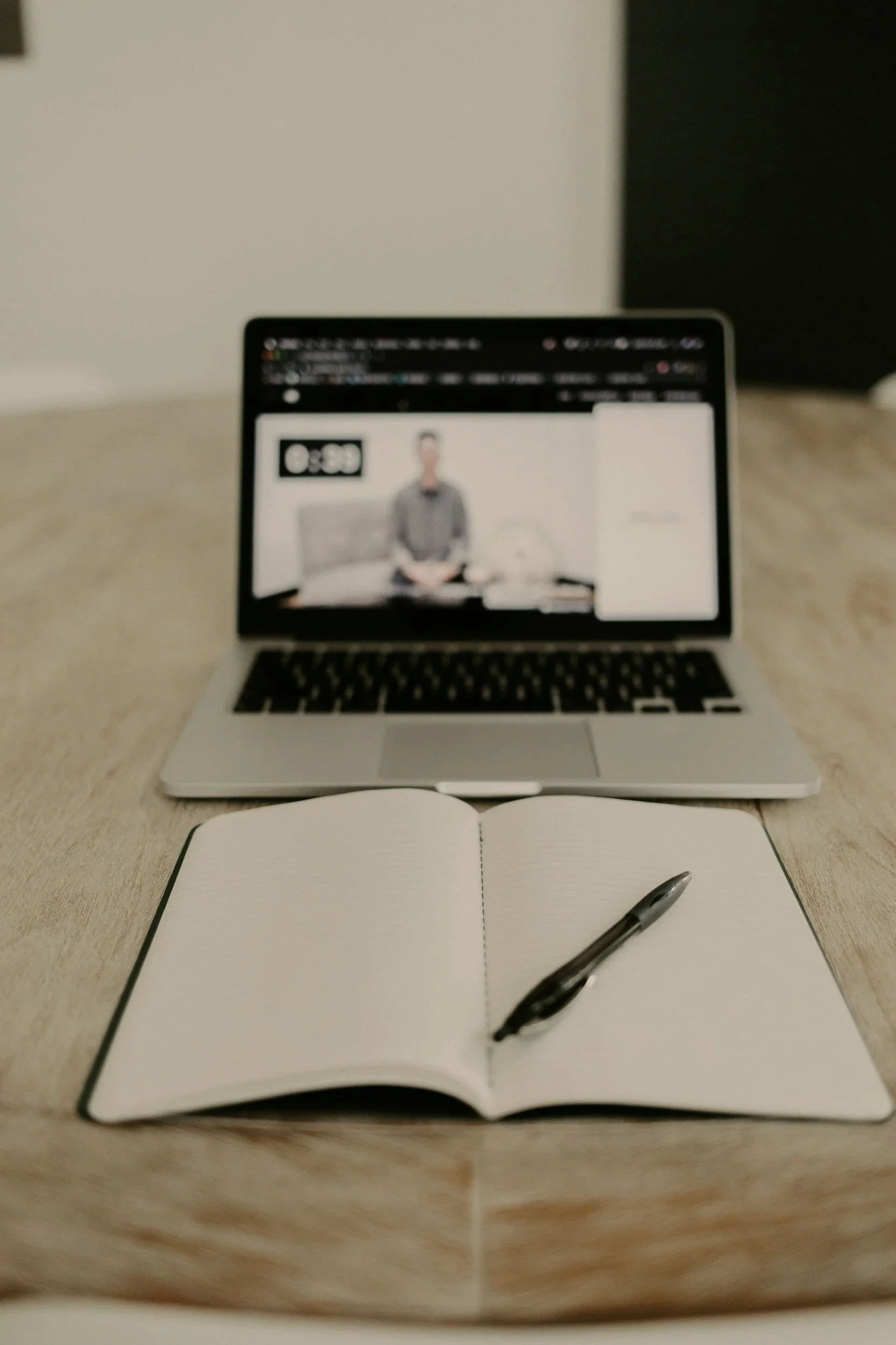 Open notebook with blank pages and pen on a wooden desk in front of a laptop displaying a blurred video of a person.