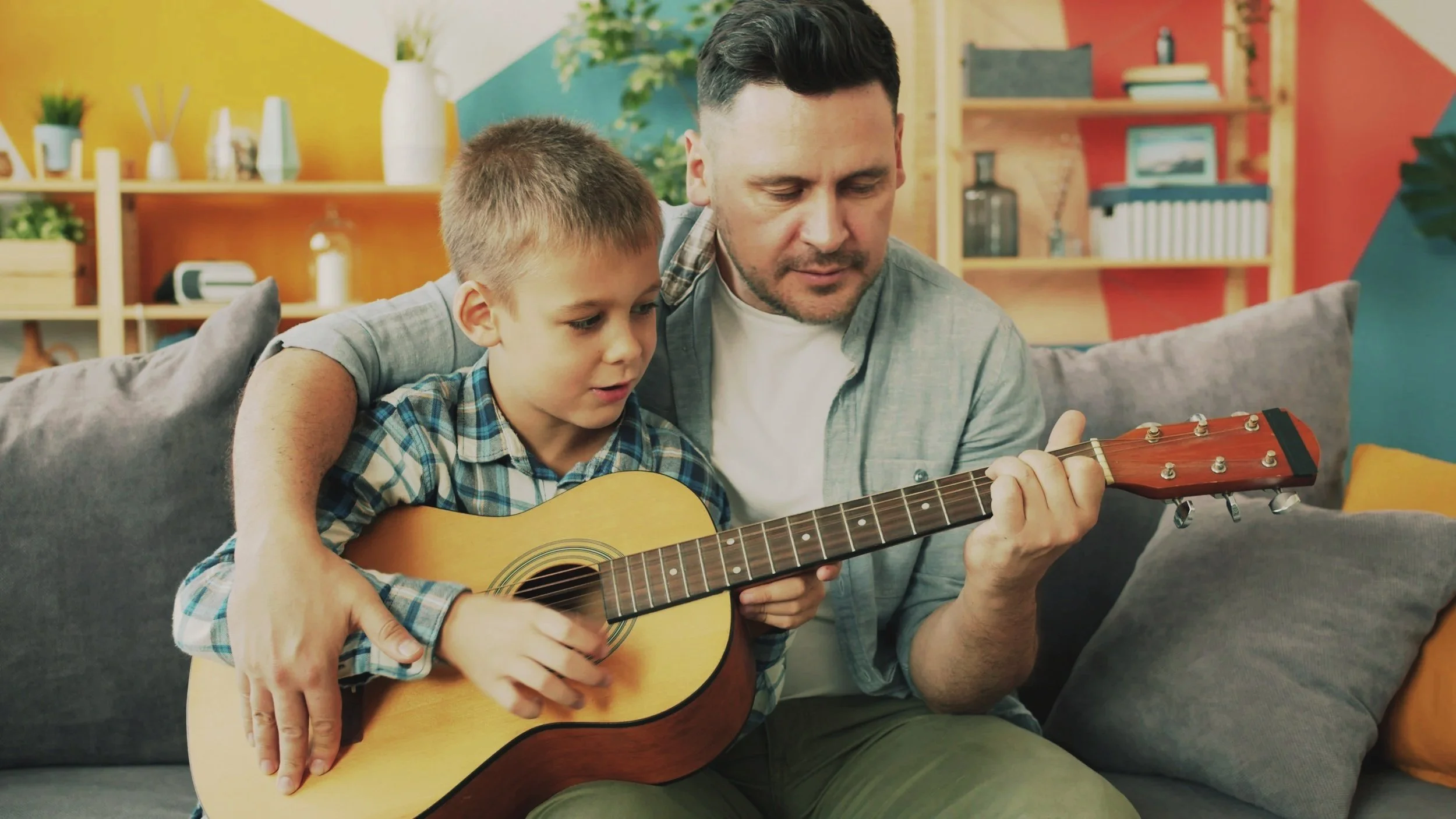 A man with dark hair and a boy with short blonde hair sit on a gray couch in a colorful room, playing a guitar together. The boy is holding the guitar and looking at it, while the man guides him. The room has a vibrant yellow, blue, and red wall with shelves holding books, plants, and decor.
