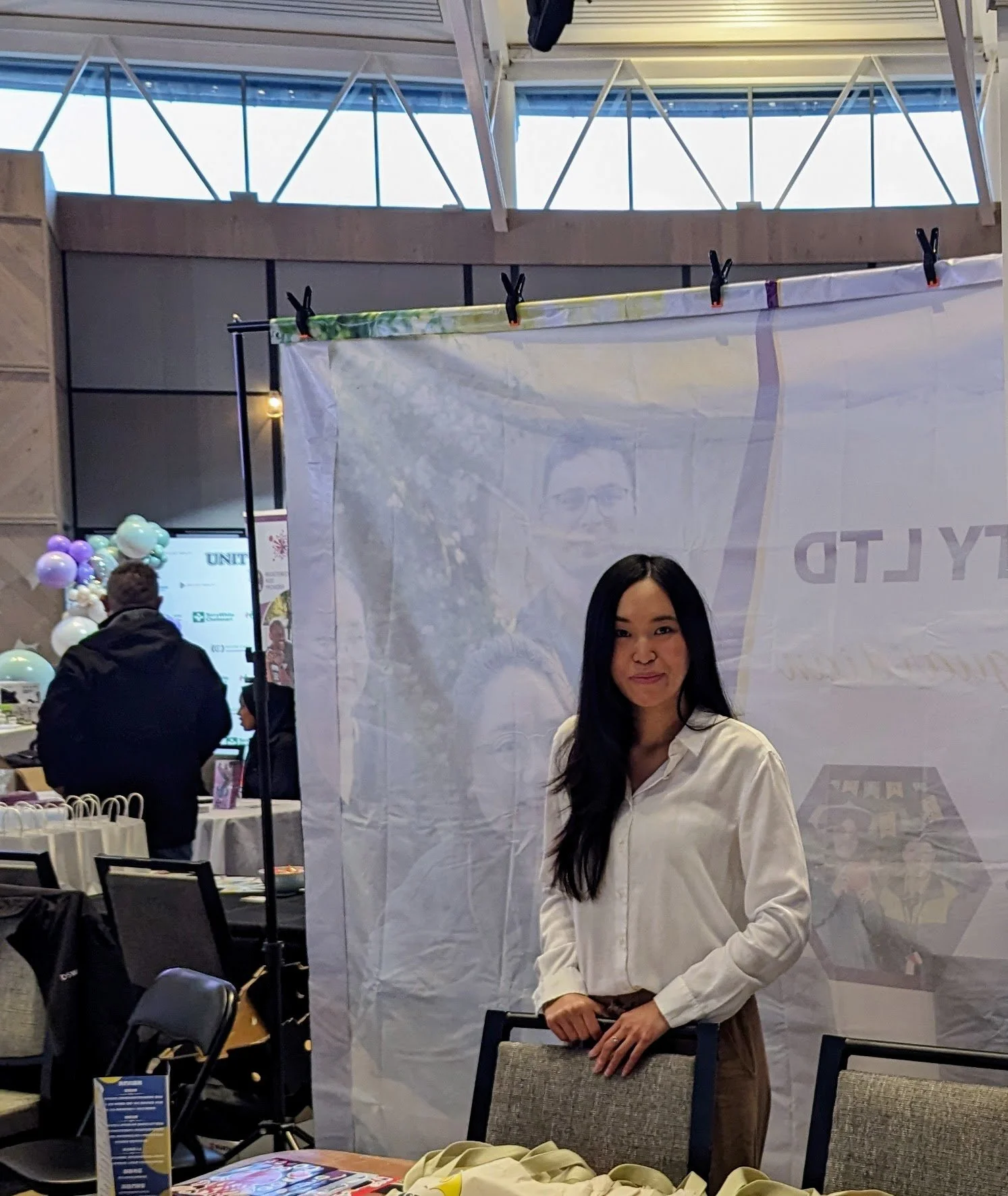 A young woman with long dark hair wearing a white blouse standing in front of a white display booth at an indoor event. There are chairs and tables nearby, and other people are present in the background.