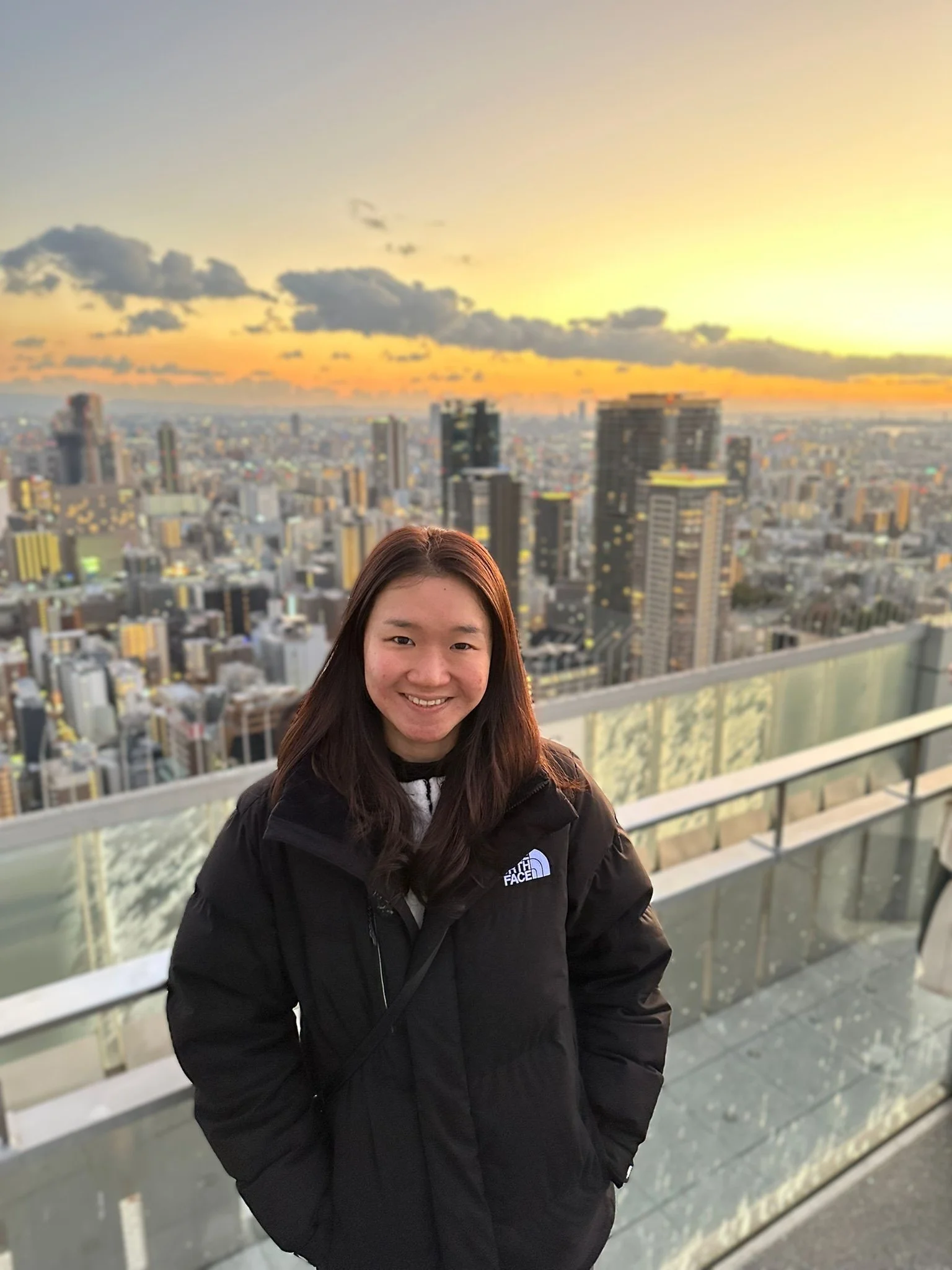 A smiling woman with long dark hair wearing a black North Face jacket standing on a balcony overlooking a cityscape at sunset.