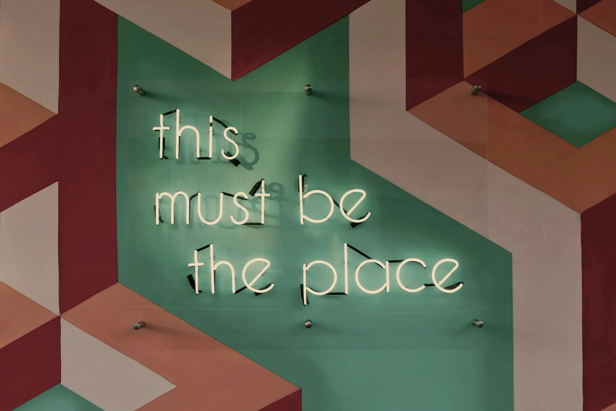Neon sign that says "this must be the place" mounted on a multi-colored geometric wall with shades of red, green, and white.