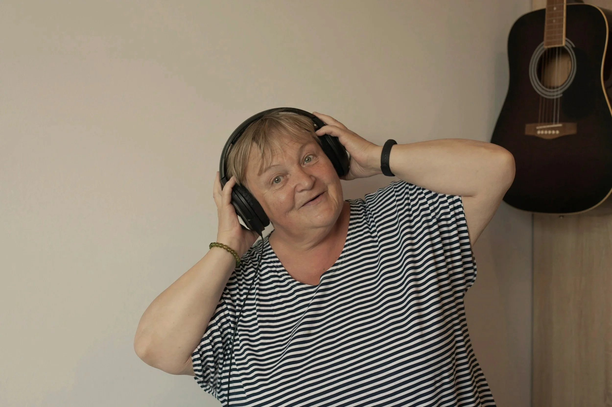 A woman with short blonde hair wearing a striped T-shirt, holding headphones on her head, smiling, standing in front of a plain wall with an acoustic guitar hanging on it.