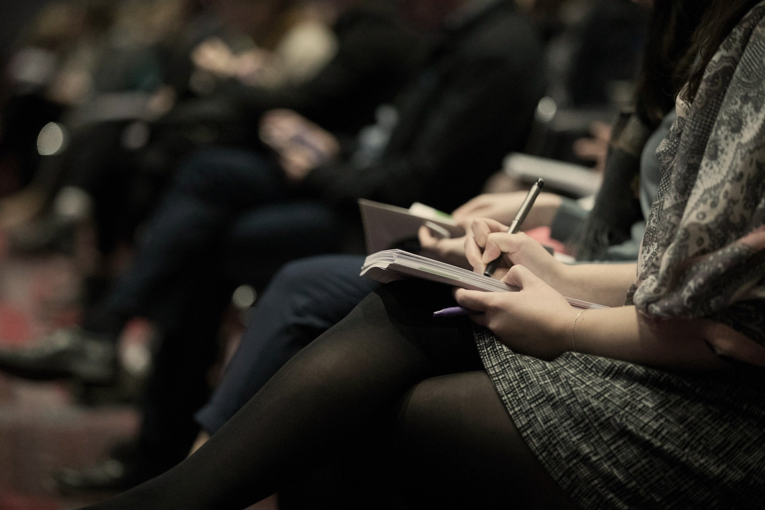 A person sitting in a crowded auditorium taking notes in a notebook with a pen.