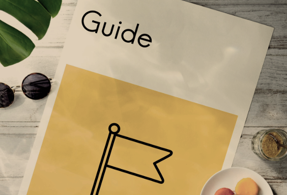 A guidebook with a simple flag illustration on the cover, placed on a wooden surface with sunglasses, a plant, and some baked goods nearby.