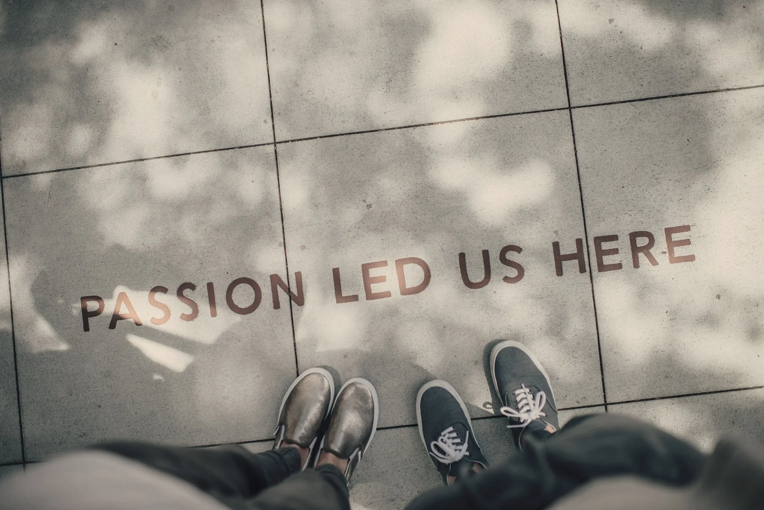 Empty tile with the words 'Passion Led Us Here' and two pairs of sneakers standing on it, seen from above, with shadows of trees cast on the tile floor.