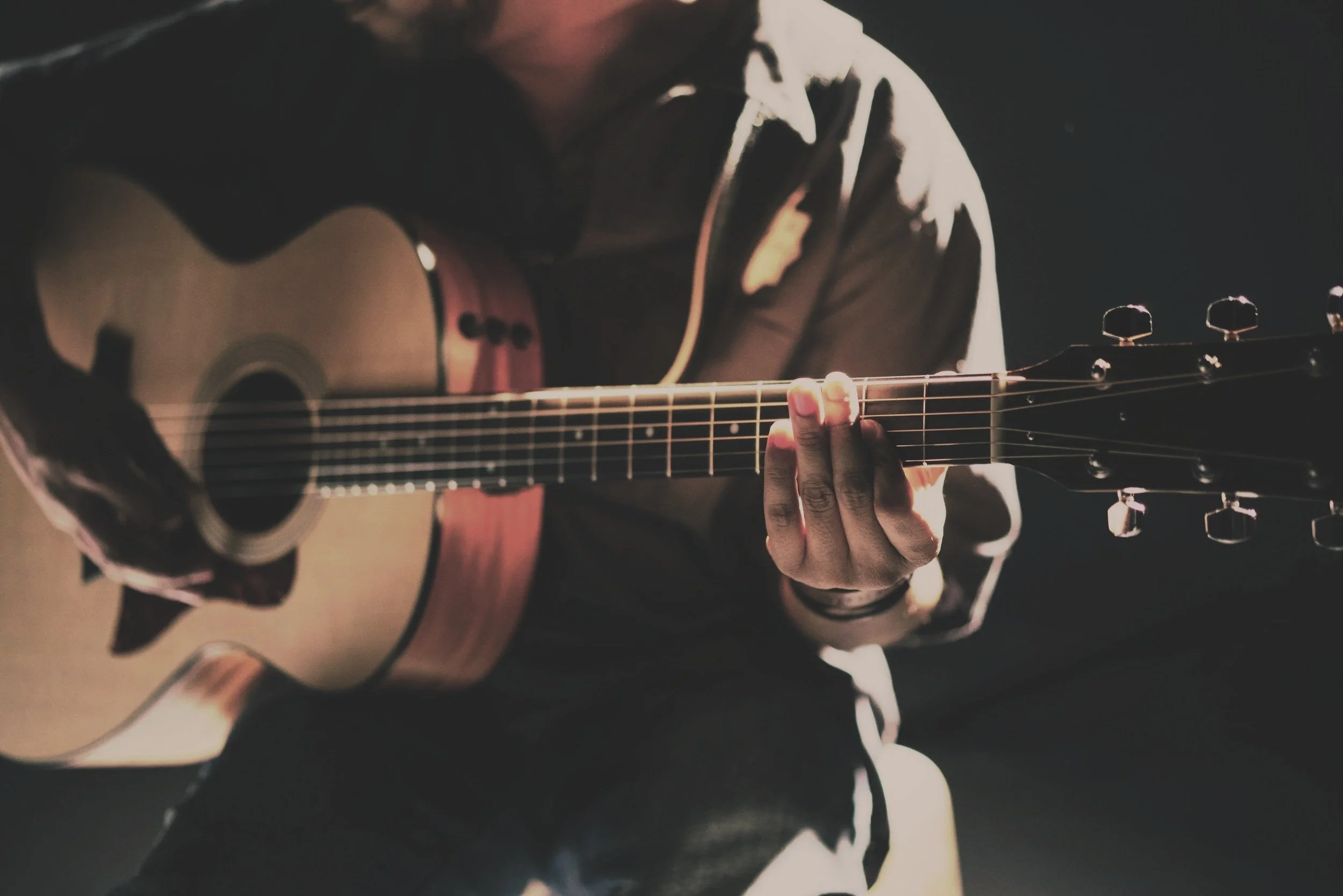 Person playing an acoustic guitar in a dimly lit setting, focusing on their hand on the strings.