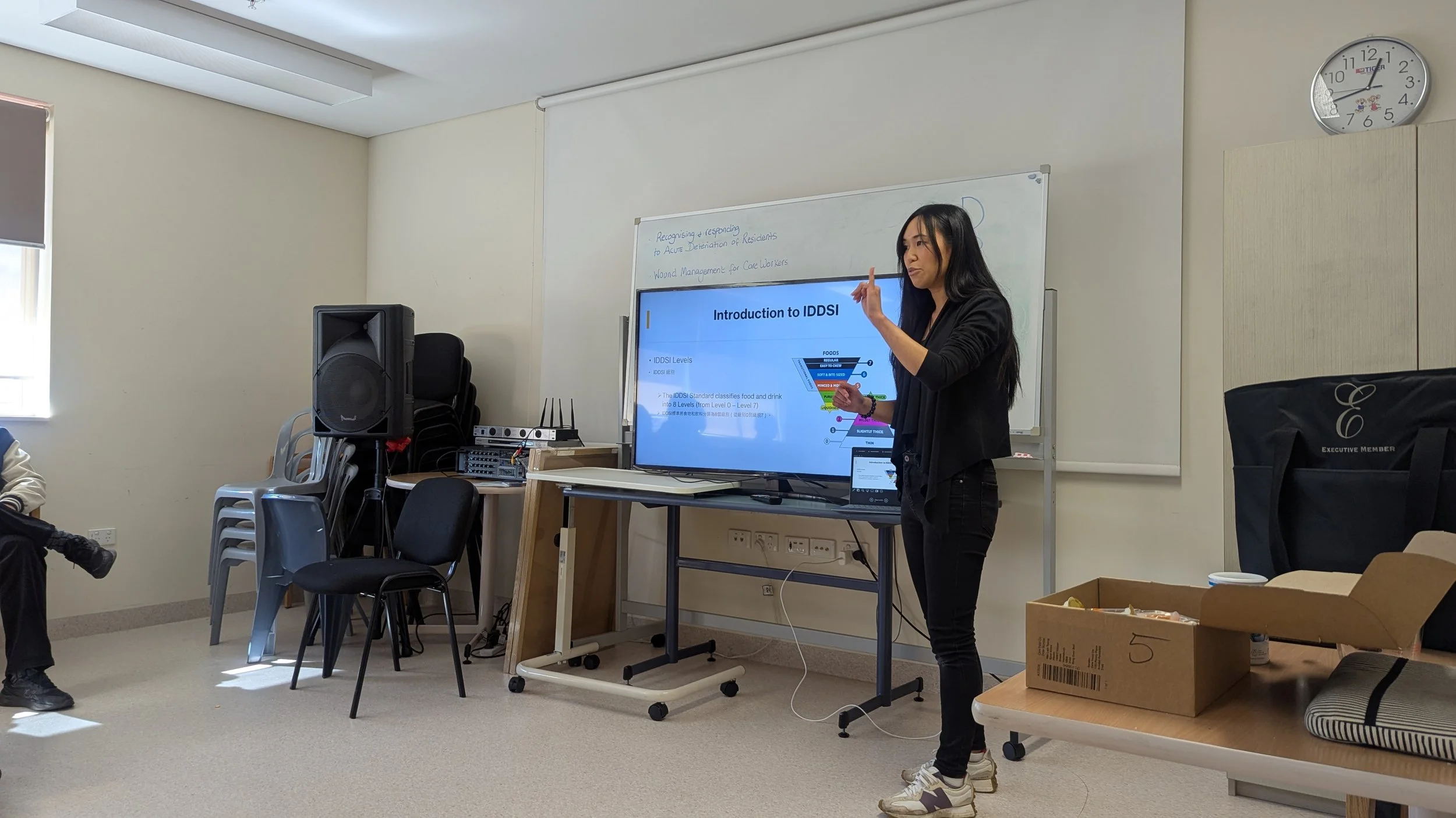 A woman giving a presentation in a classroom, standing in front of a large screen displaying a slide titled 'Introduction to IDDSI'.