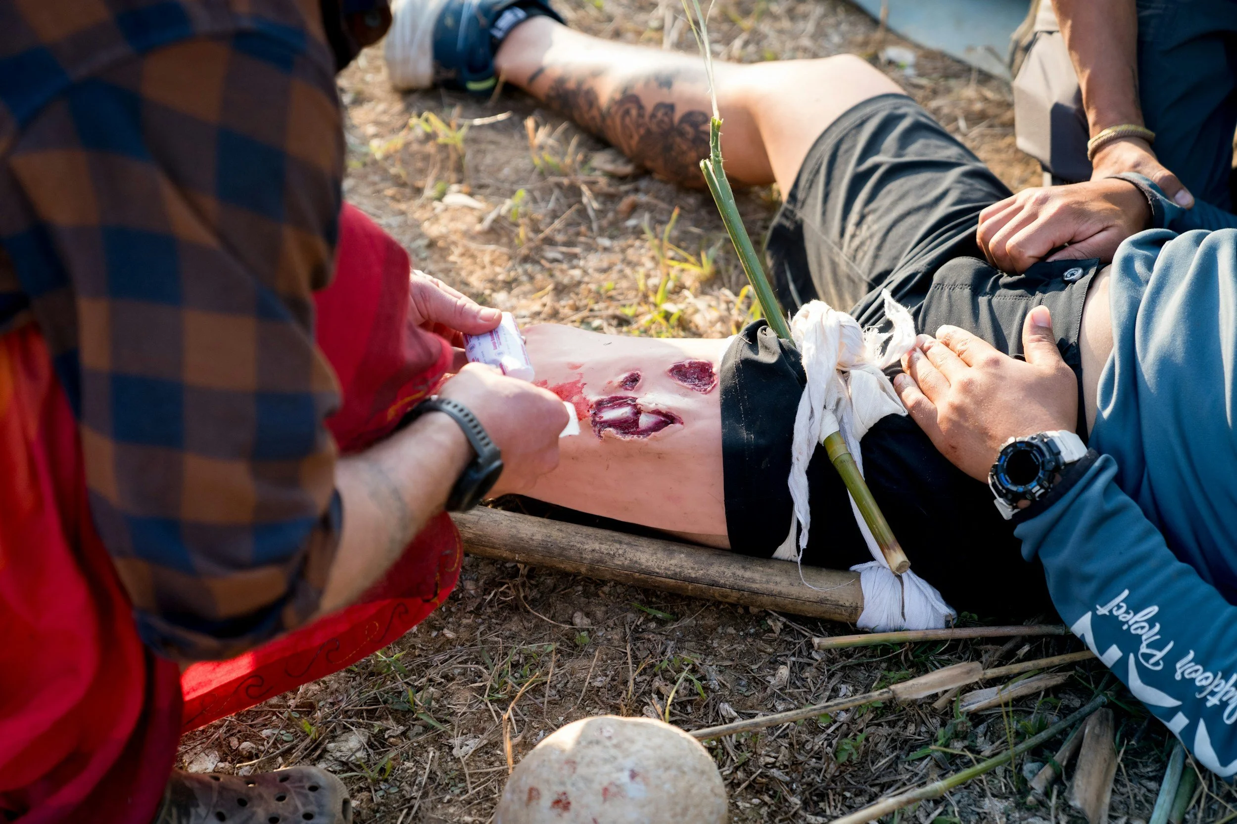 Two individuals are assisting a person with a leg injury outdoors on rocky ground. The injured person is lying on a wooden surface with blood and an open wound on the thigh. One person is applying a bandage or dressing, while the other is holding the leg. The scene appears to be an emergency medical response.