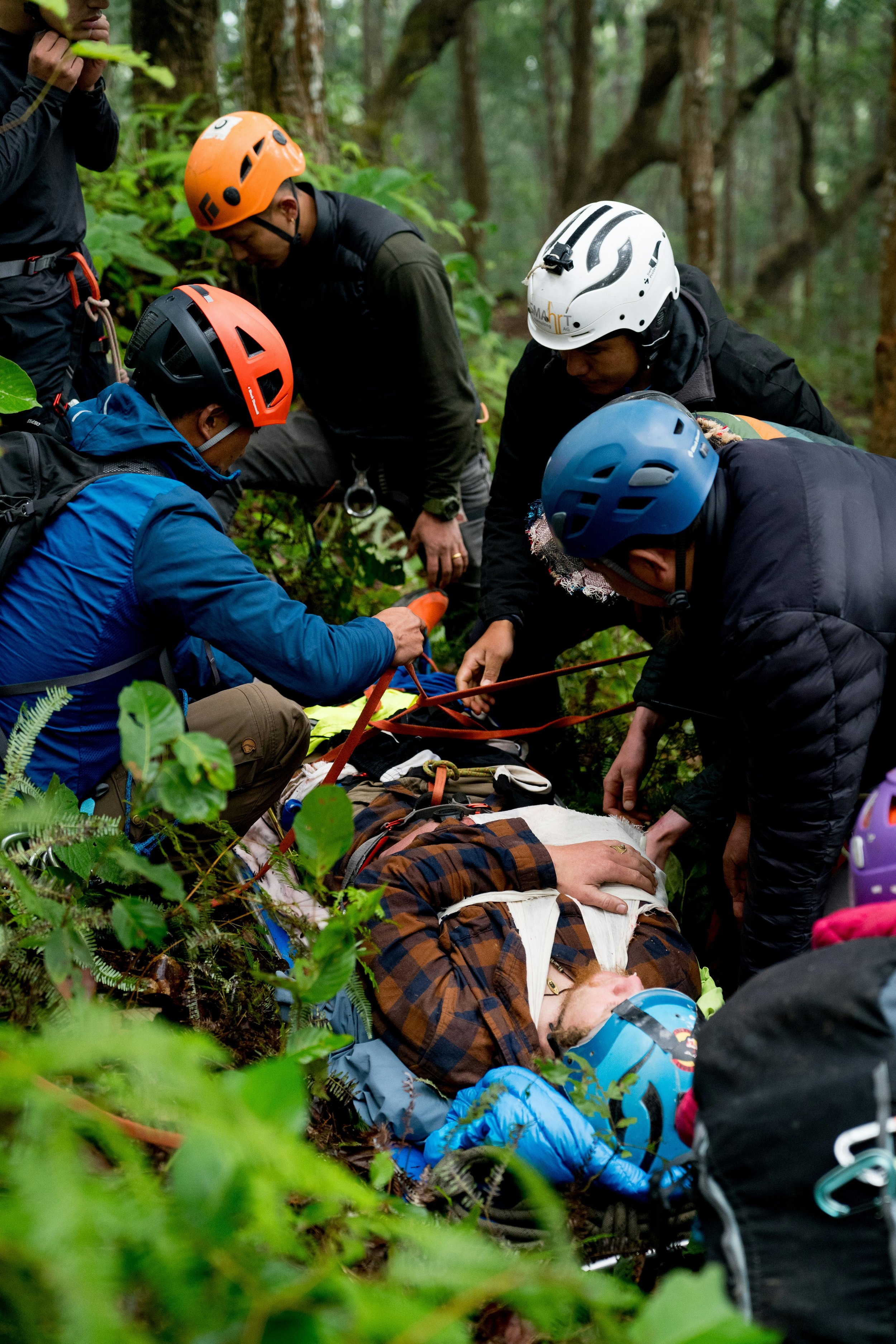 Rescue team attending to an injured person lying on the forest ground, with multiple rescuers wearing helmets and outdoor gear, surrounded by dense green foliage.