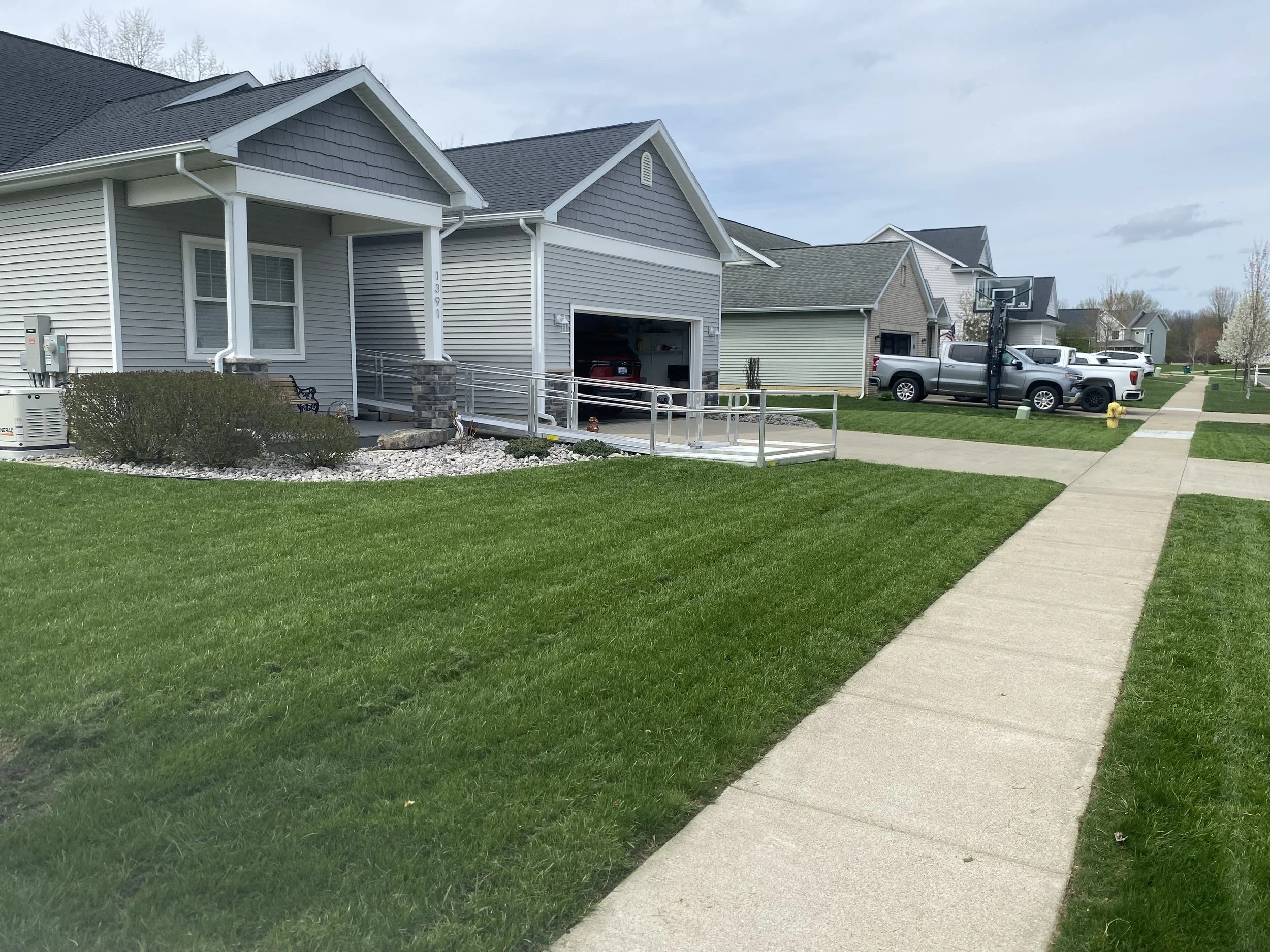 A suburban neighborhood scene with a sidewalk, green lawn, and multiple houses with driveways. The houses have light-colored siding and dark roofing, with parked trucks and a basketball hoop visible. The sky is partly cloudy.