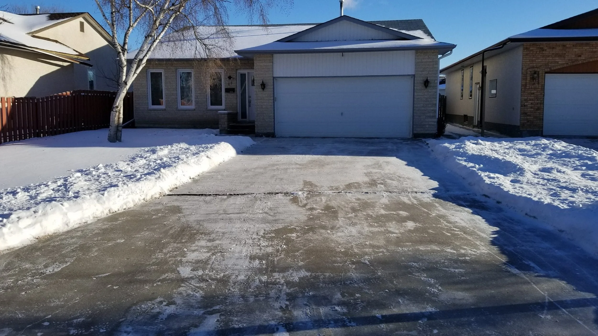A suburban house with a white garage door, snow-covered driveway, a leafless tree in front, and neighboring homes on either side, during daytime with clear blue skies.