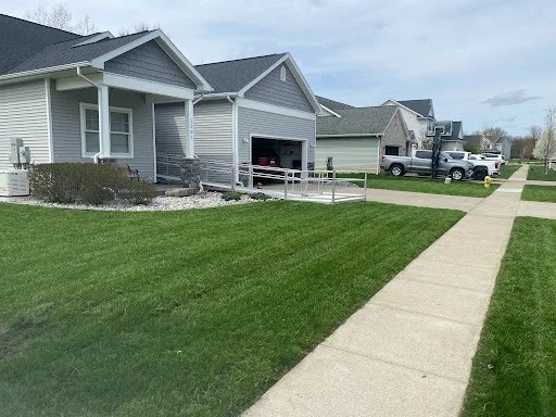 Neighborhood street with single-family houses, green lawns, cars parked in driveways, and a clear blue sky.