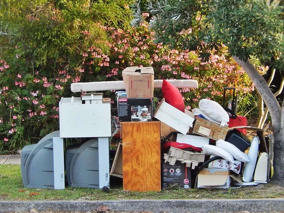 A pile of discarded household items and boxes on the sidewalk next to a grassy patch with pink flowering bushes and trees.