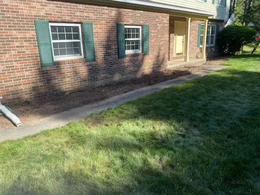 A brick house with two windows featuring green shutters, a yellow door, and a small front yard with a sidewalk and green grass.