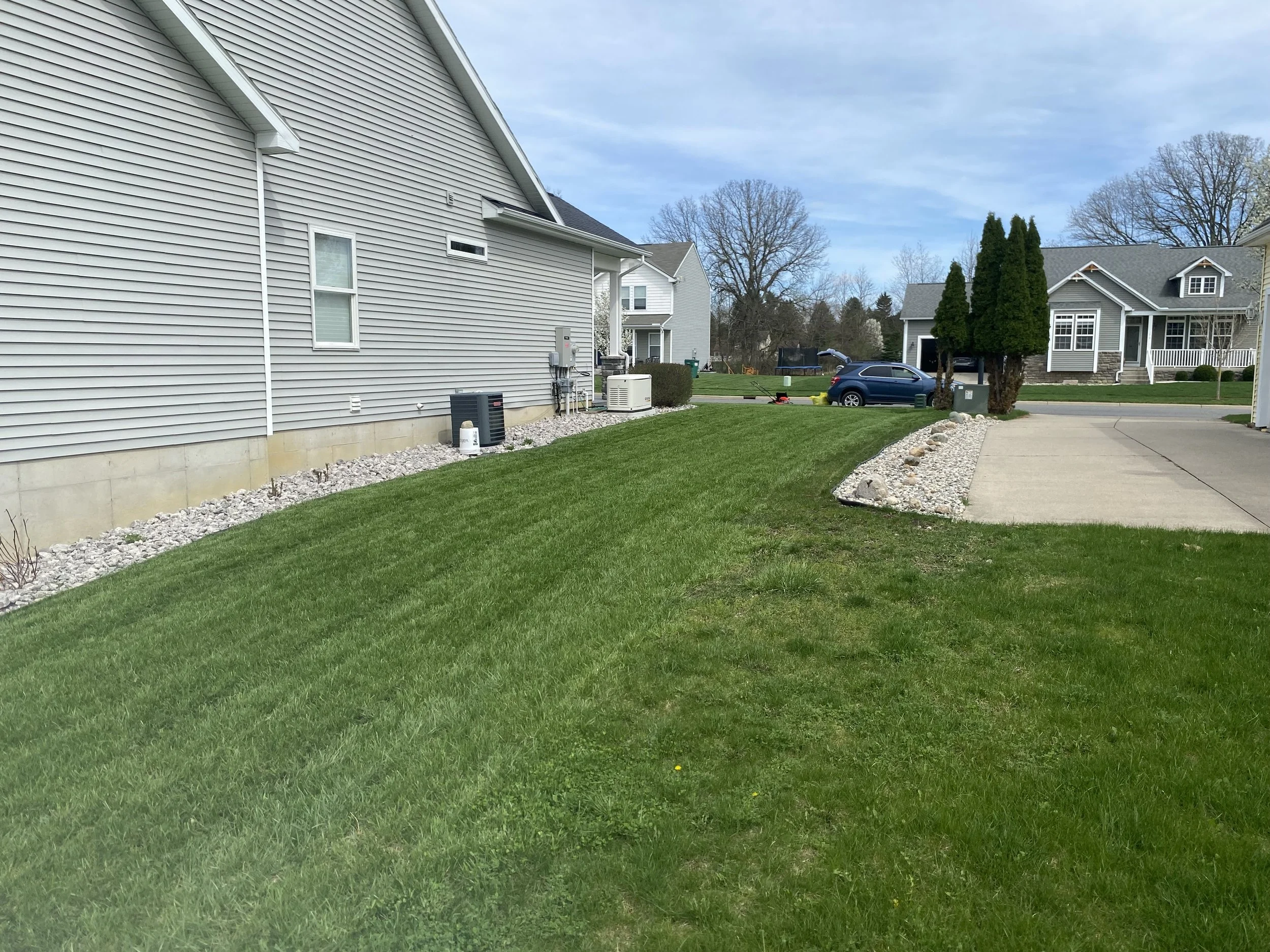 A suburban neighborhood with green lawns, houses with vinyl siding, a driveway, trees, and a blue sky.