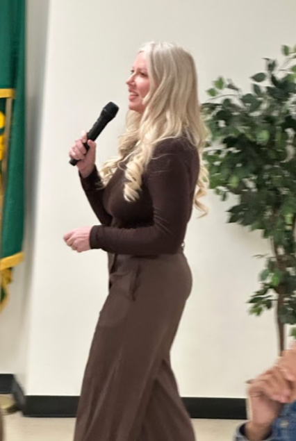 A woman with long, curly blonde hair holding a microphone and speaking at an indoor event, standing next to a potted plant.