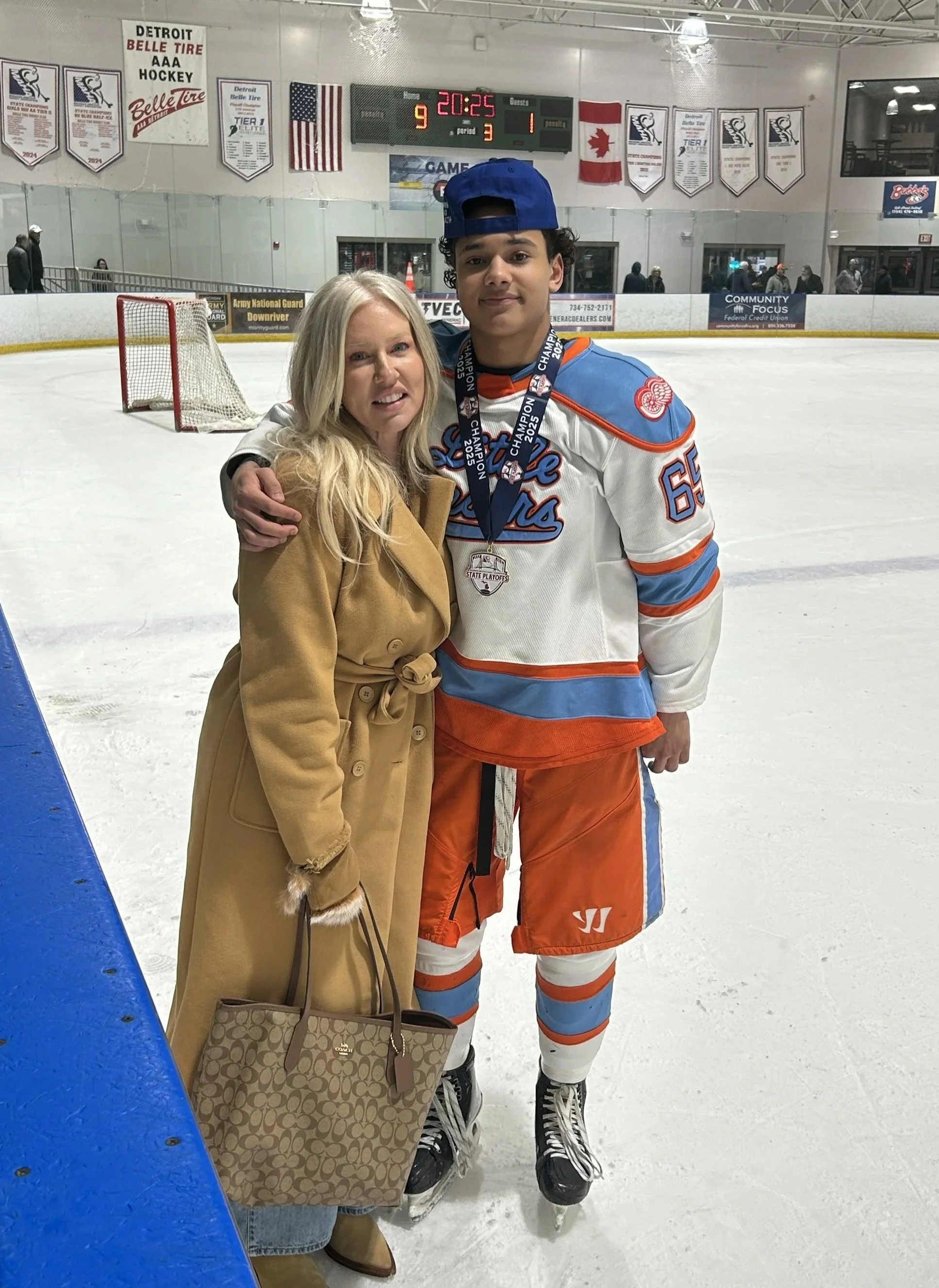 A woman and a young male hockey player posing together inside an ice rink. The hockey player is wearing an orange and blue Detroit Lakes jersey and a medal. The woman is wearing a tan coat and holding a designer handbag. The scoreboard shows the game ended with Detroit Lakes winning 9-1.