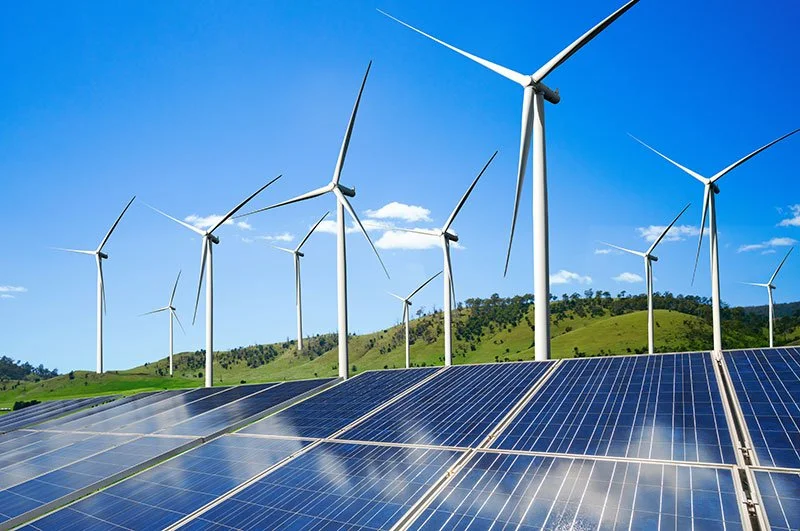 Wind turbines and solar panels on a sunny day with a hill in the background.