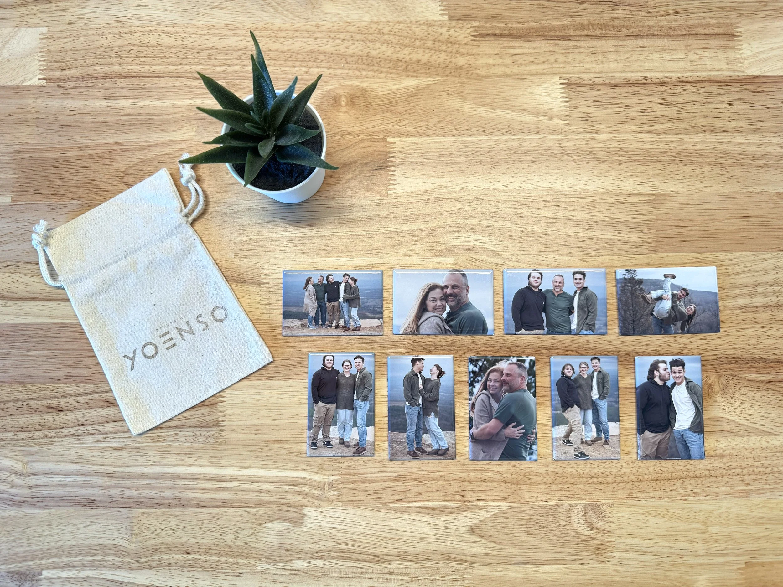 Collection of eight printed photographs of a group of people displaying friends and family, arranged on a wooden surface near a small potted succulent plant and a cloth bag labeled 'YONO'.