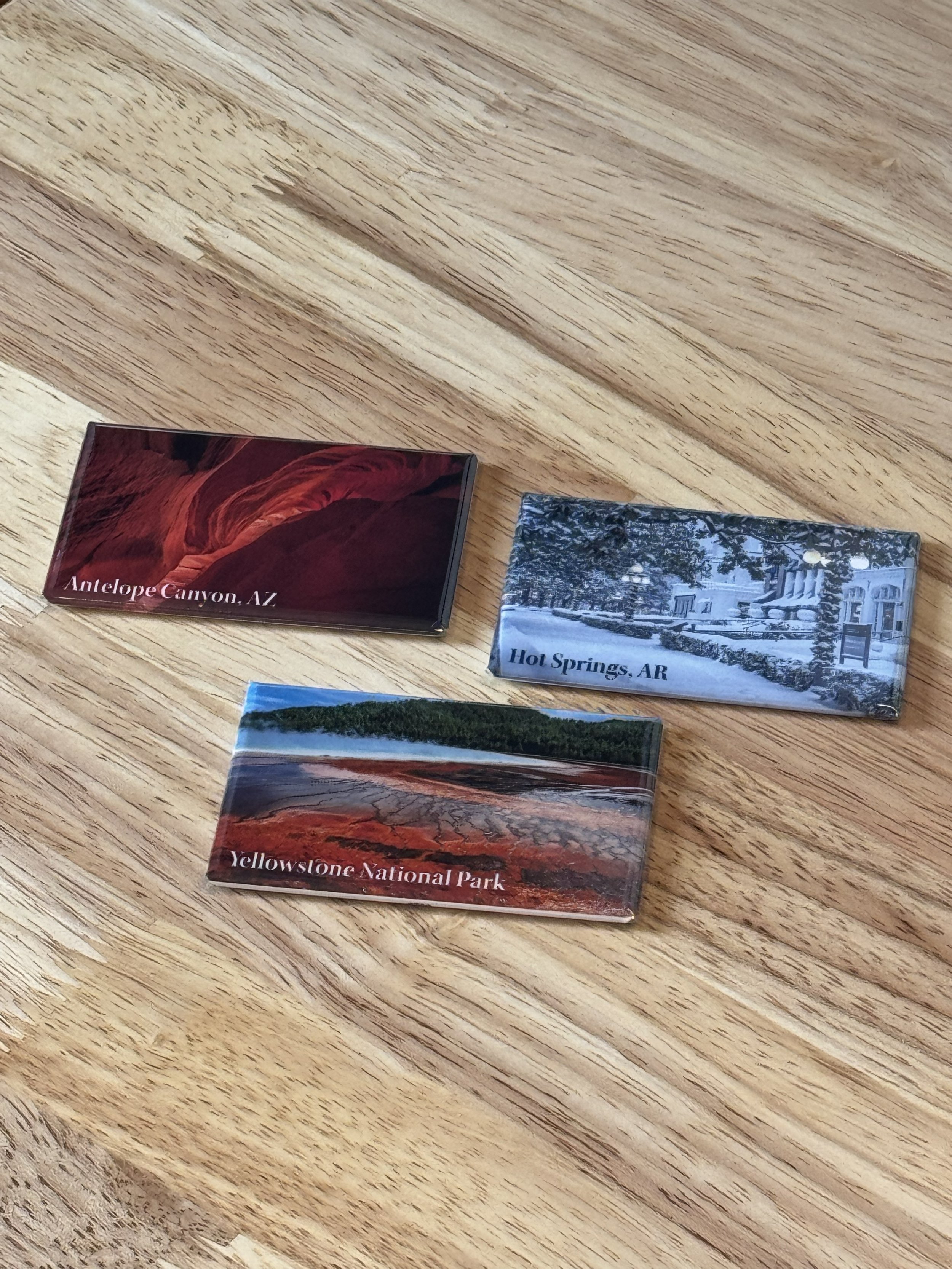 Three souvenir magnets on a wooden surface, each featuring a different location: Antelope Canyon, Arizona; Hot Springs, Arkansas; Yellowstone National Park.
