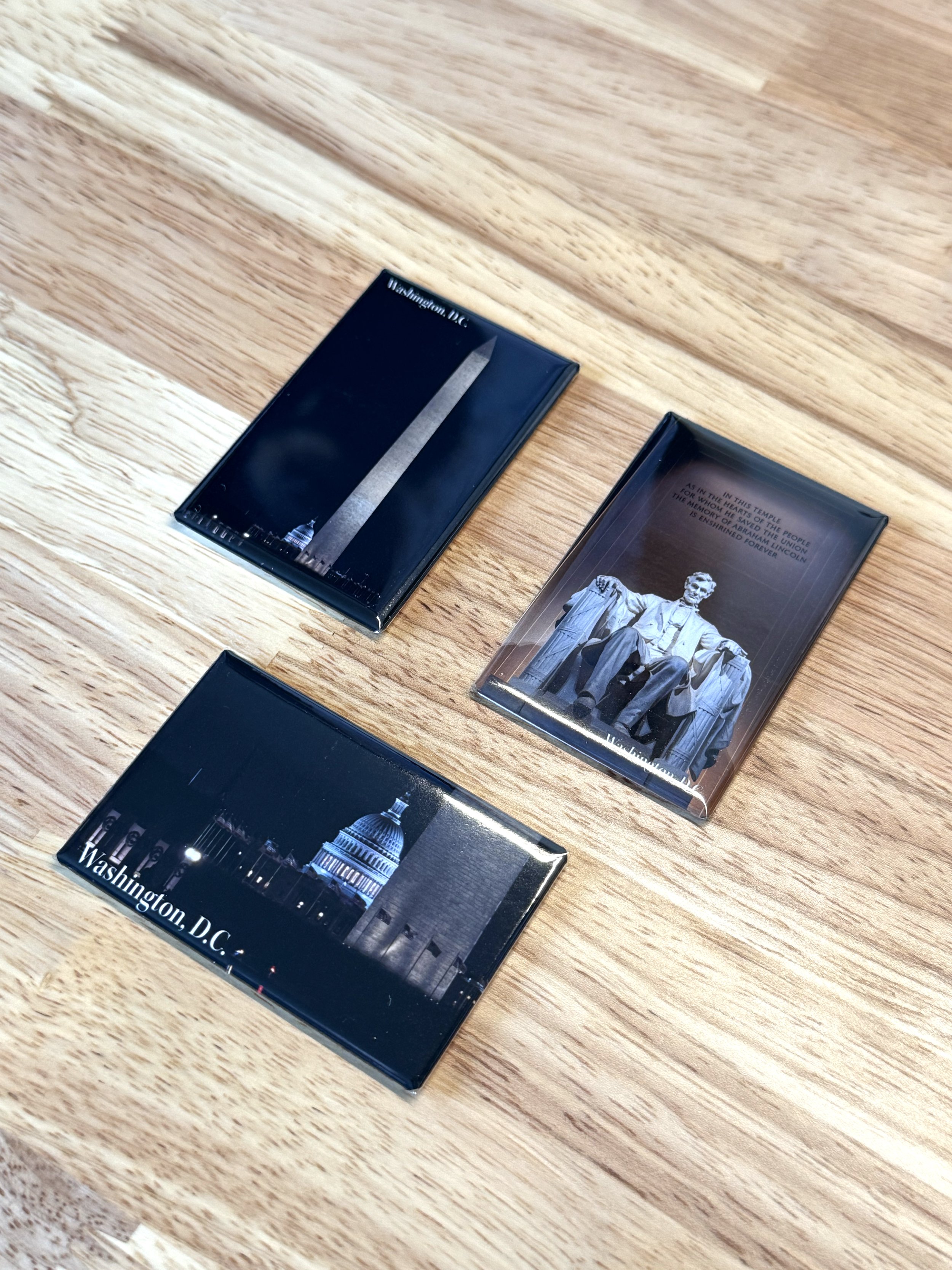Three photo magnets featuring Washington, D.C. landmarks and monuments, including the Washington Monument, the Lincoln Memorial, and the Capitol Building, placed on a wooden surface.