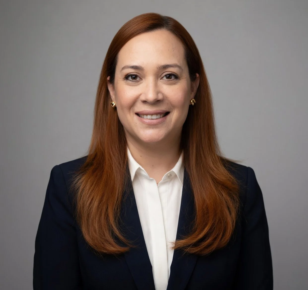 Professional headshot of a woman with long red hair, wearing a navy blazer and white blouse, smiling at the camera against a gray background.