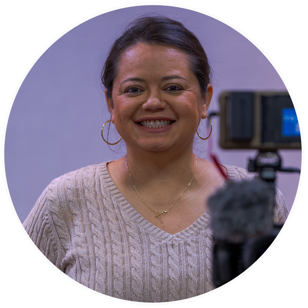 A woman smiling in front of a camera, wearing a beige sweater, hoop earrings, and gold jewelry.