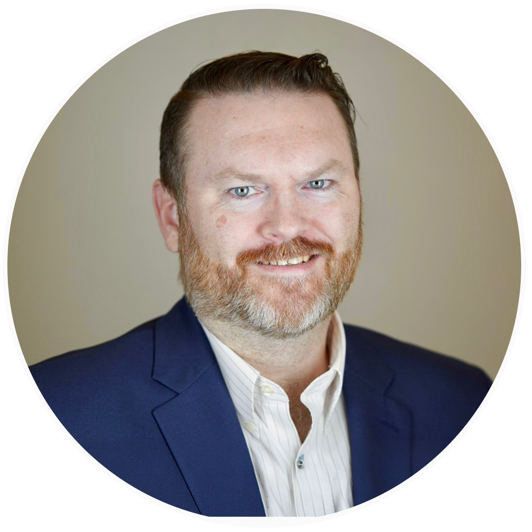 Headshot of a smiling man with a reddish beard, wearing a dark blue suit and a white collared shirt, against a beige background.