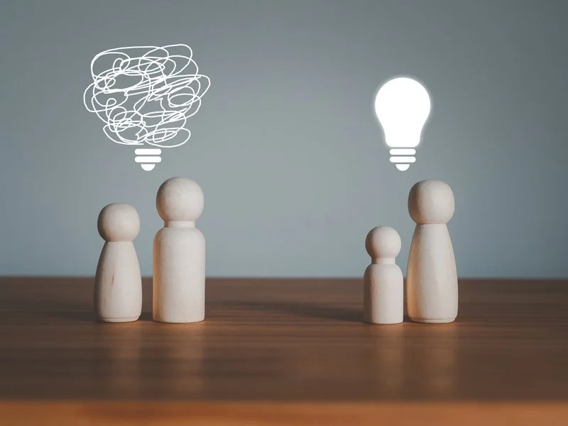 Four wooden figurines on a wooden surface with digital lightbulb and scribbled cloud icons above them, symbolizing ideas and thinking.