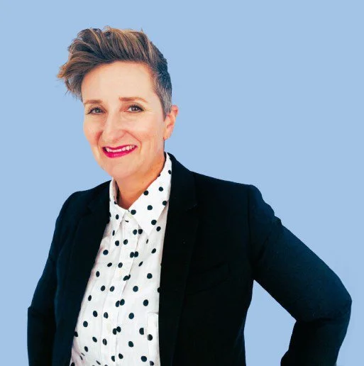 A woman with short, styled hair smiling, dressed in a polka dot shirt and a dark blazer, standing against a light blue background.