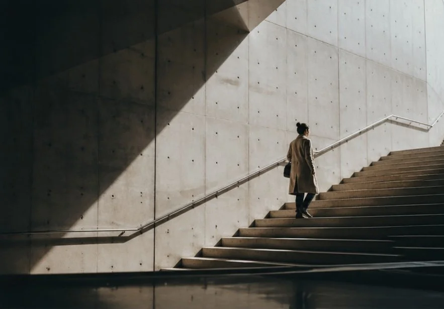 A woman in a long coat walking up a staircase against a concrete wall in a modern building.