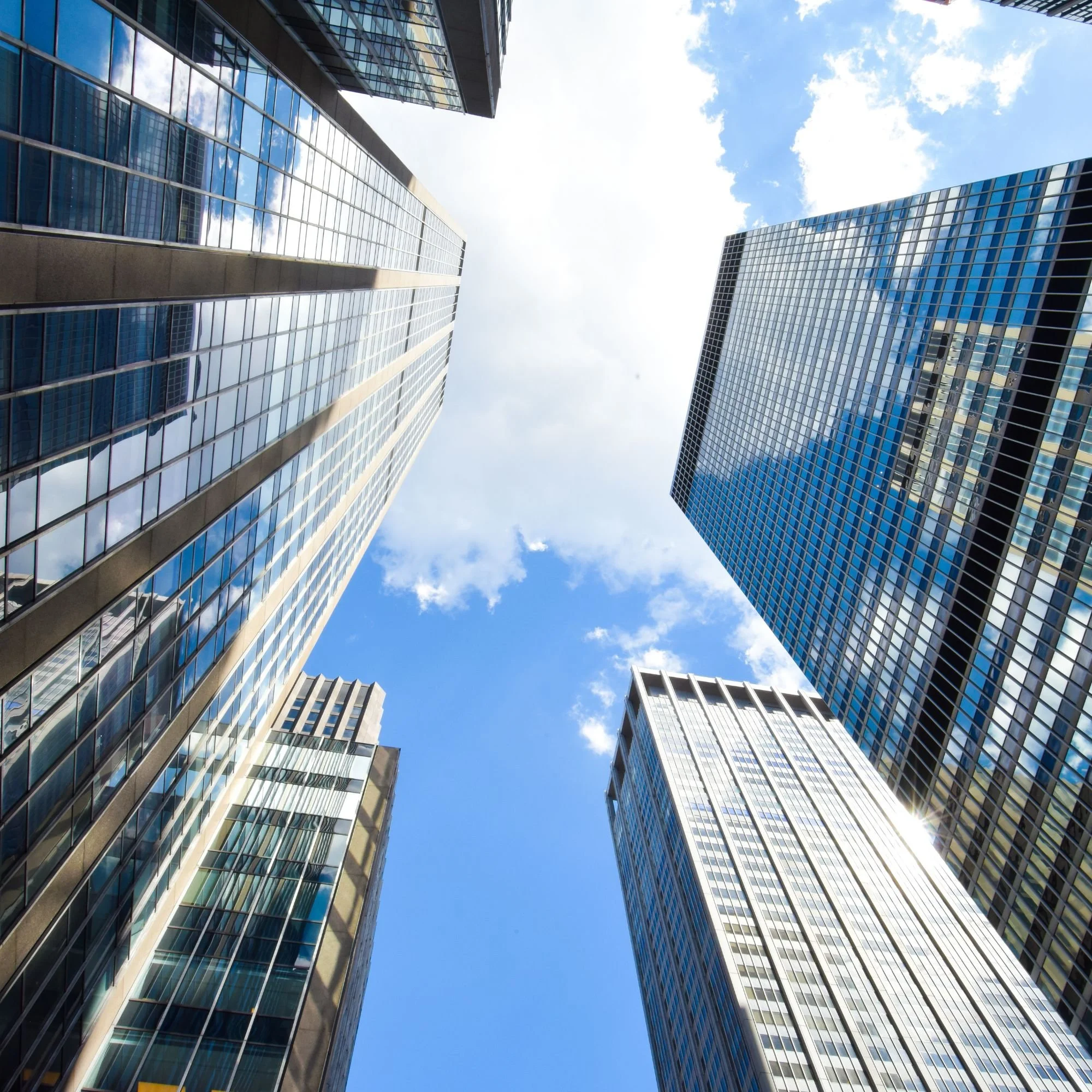 Looking upward at tall glass skyscrapers with reflective windows against a bright blue sky with scattered clouds.