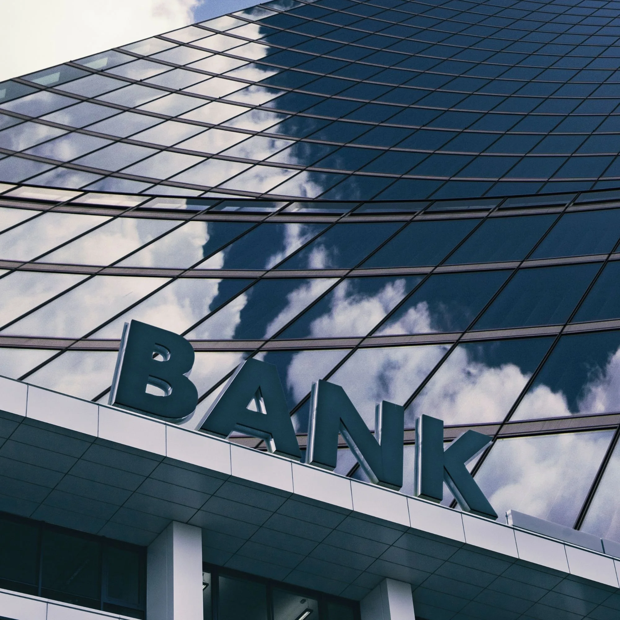 Low-angle view of a modern glass office building with reflective windows, the sky and clouds are reflected in the windows, and part of a large letter B sign is visible at the bottom of the image.
