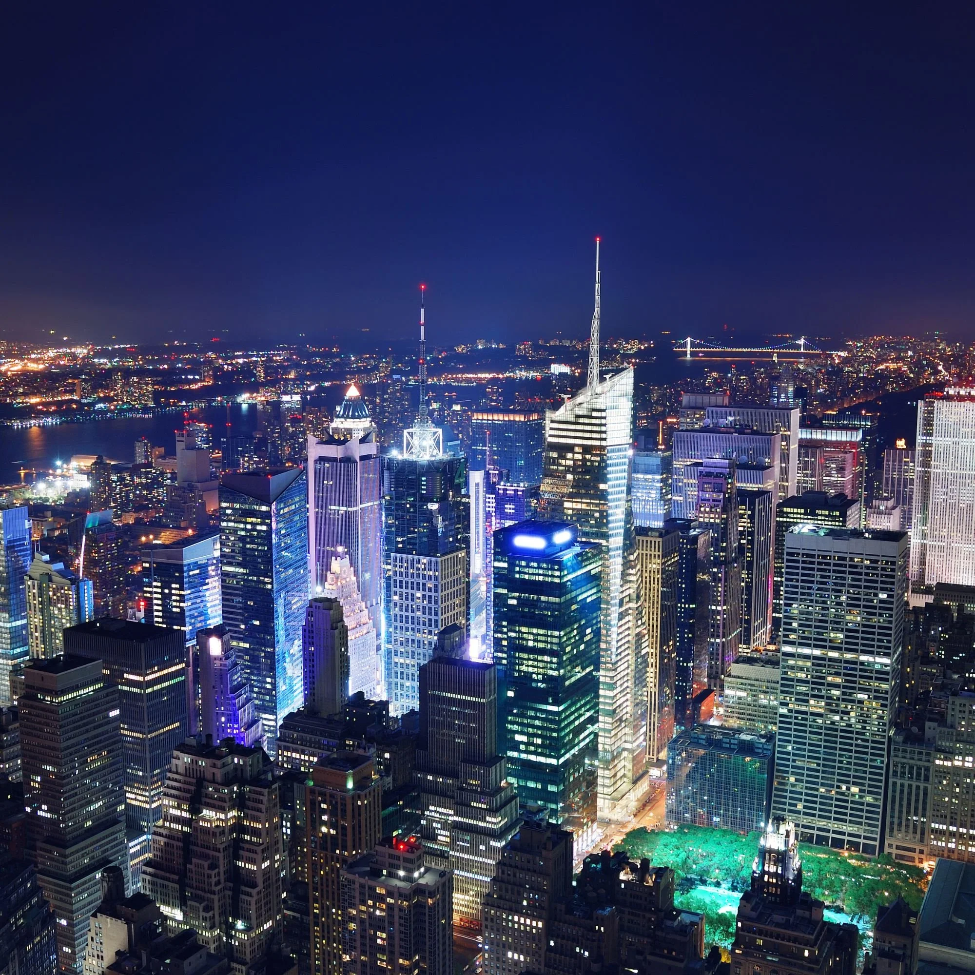 Nighttime city skyline with illuminated skyscrapers, a river, and a bridge in the background.