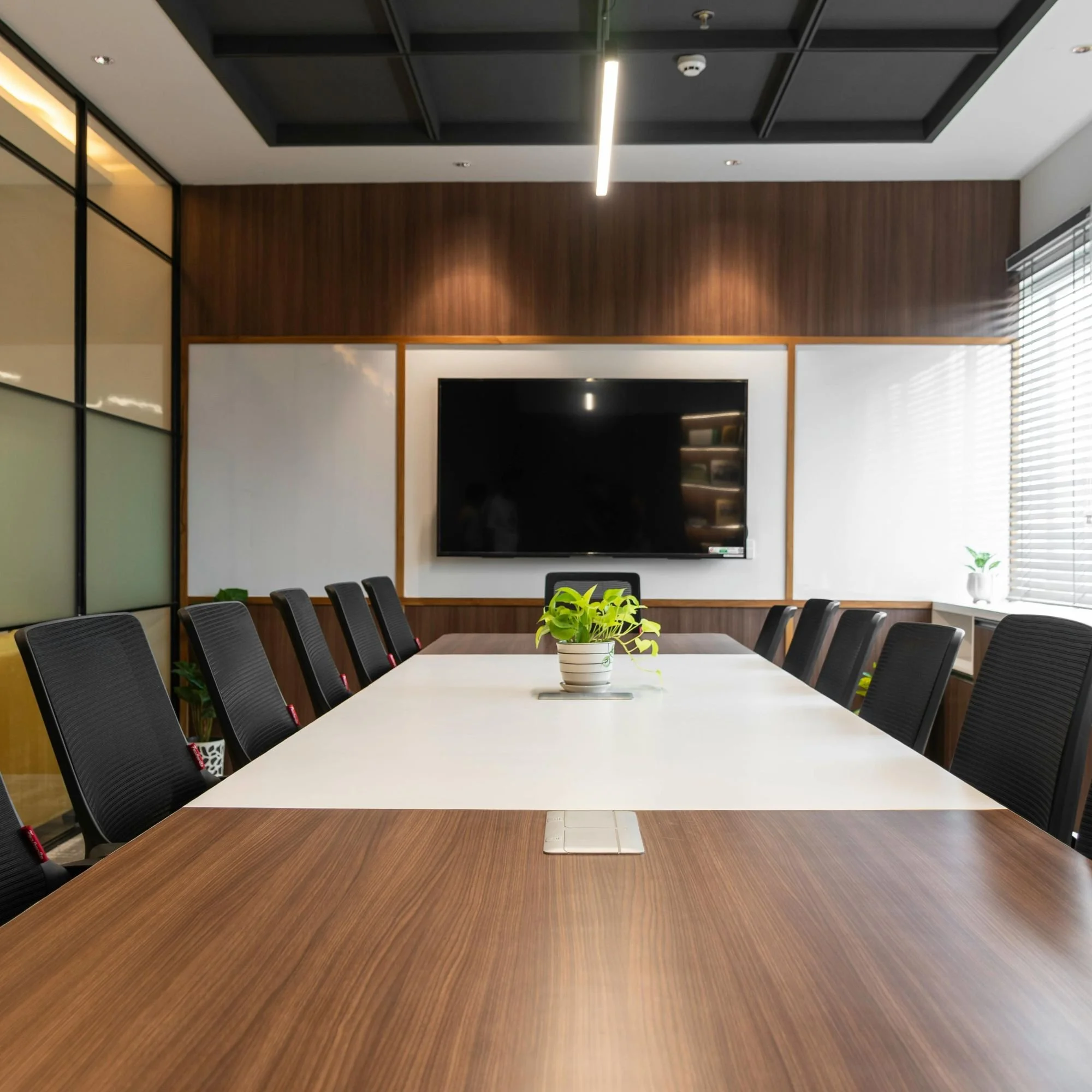 A modern conference room with a large wooden table surrounded by black chairs. On the wall, there is a large flat-screen TV, and a potted plant is placed on the table. The room has a wooden accent wall, a window with blinds, and ceiling lights.