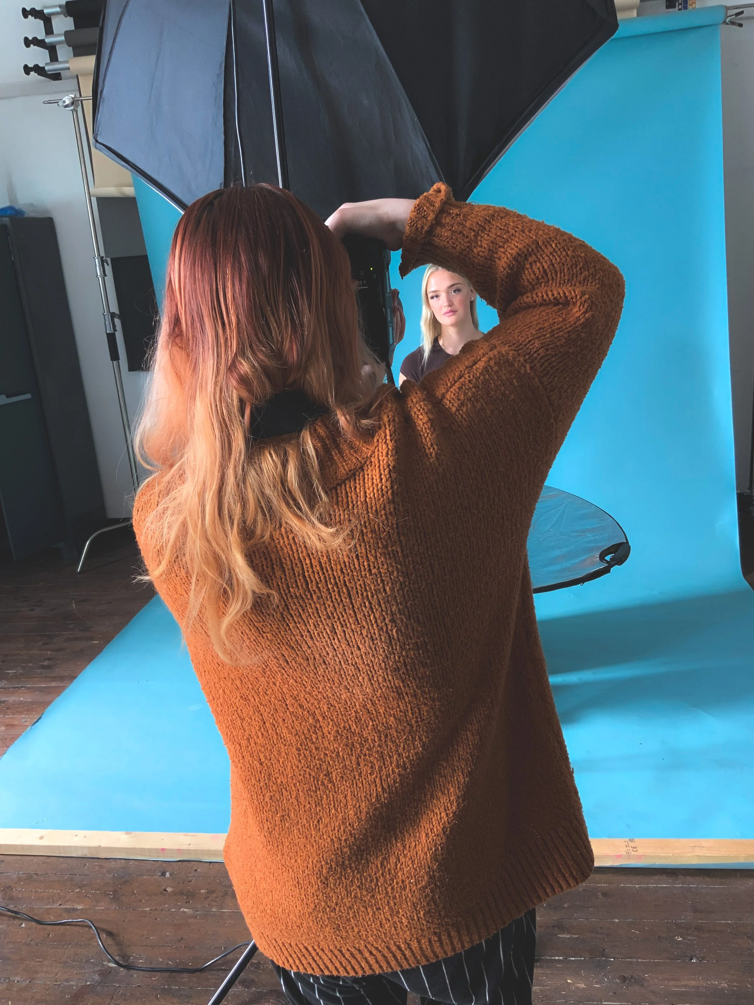 A woman with long red hair wearing a brown sweater is taking a photograph of a blonde woman with a black shirt against a blue background in a photo studio.
