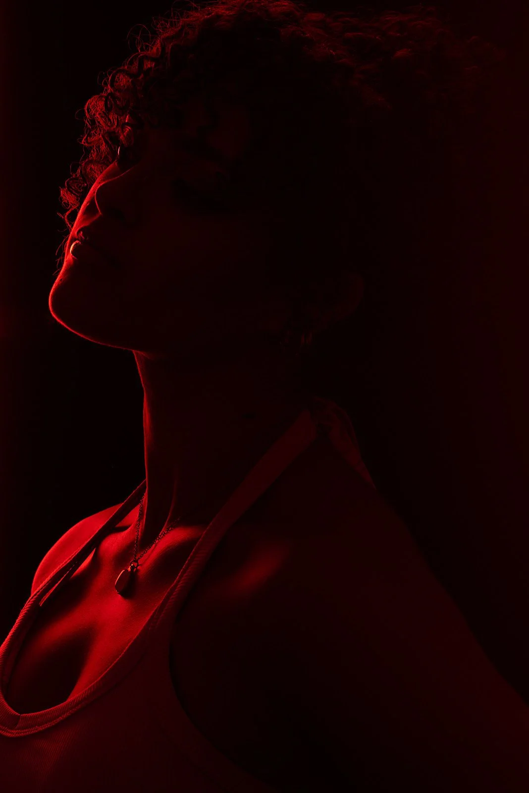 Silhouette of a woman with curly hair wearing a tank top and a necklace, illuminated by red light against a dark background.