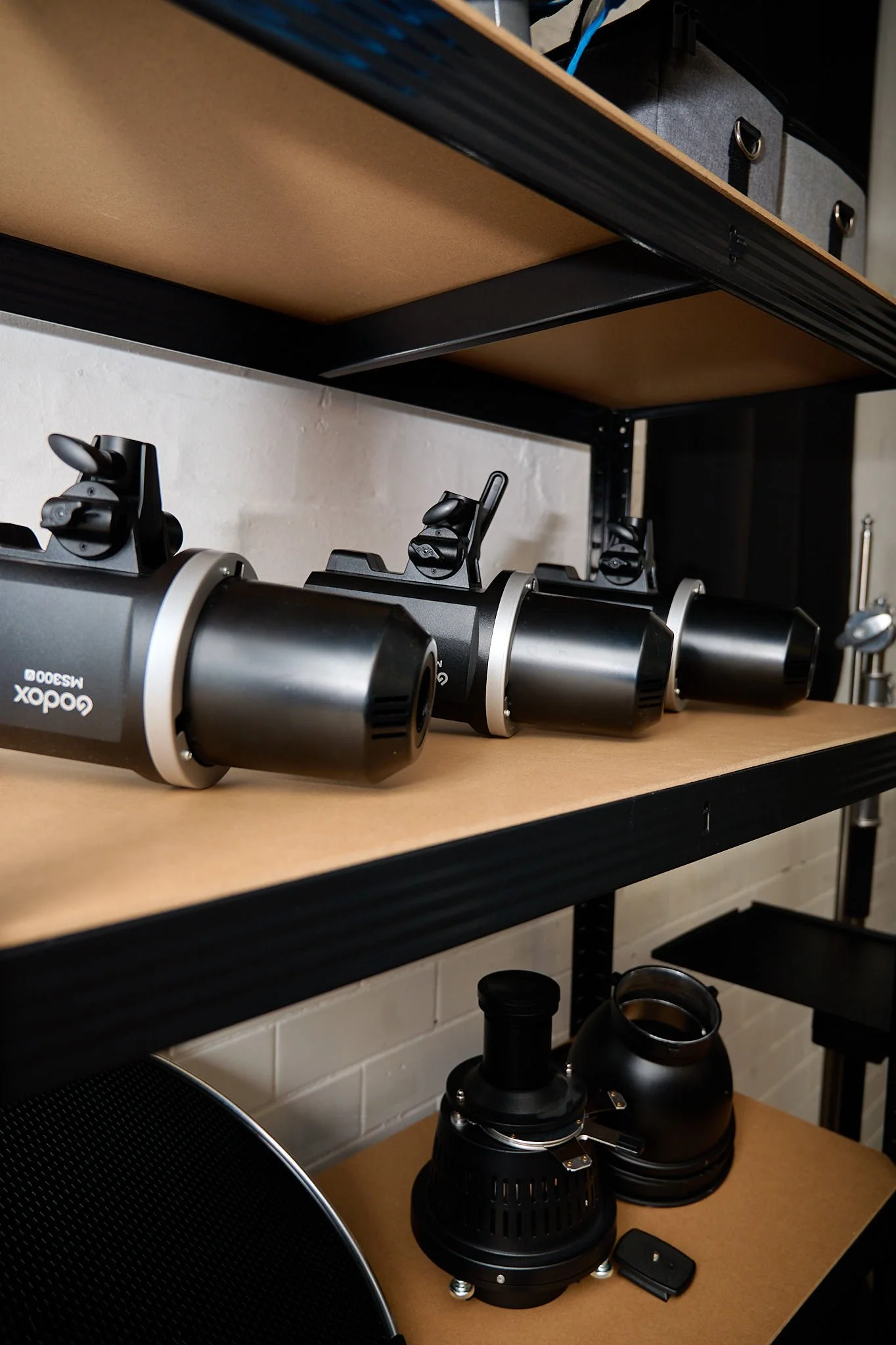 A microscope on a wooden desk below a metal and wood shelf that holds a black storage box and other equipment.